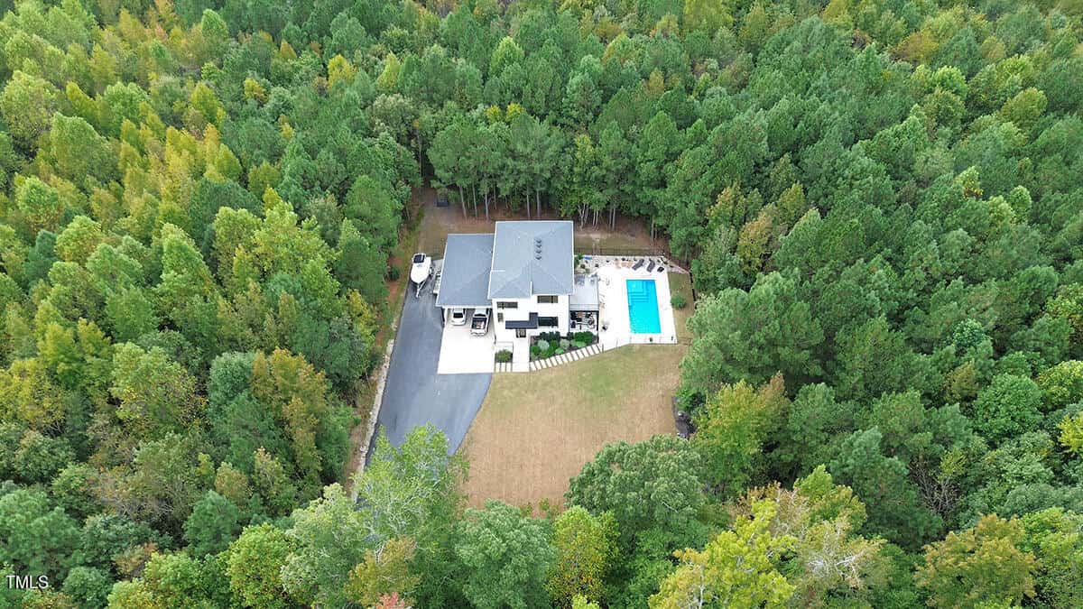 Aerial view of a modern house surrounded by dense green trees, with a driveway, a lawn, and a rectangular swimming pool in the backyard. The property is nestled in a forested area.