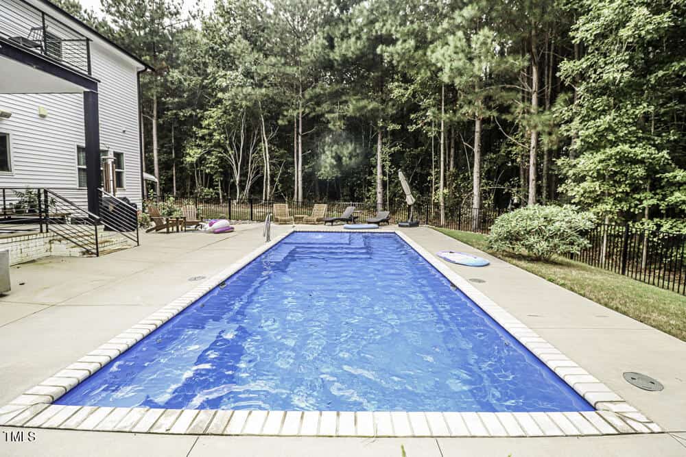 A rectangular outdoor swimming pool with a paved deck, lounge chairs, and floaties, surrounded by trees and greenery, next to a modern house.