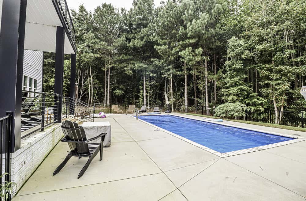 Backyard with a rectangular swimming pool, lounge chairs, patio furniture, and a forest of tall trees in the background. A covered porch is on the left side of the image.