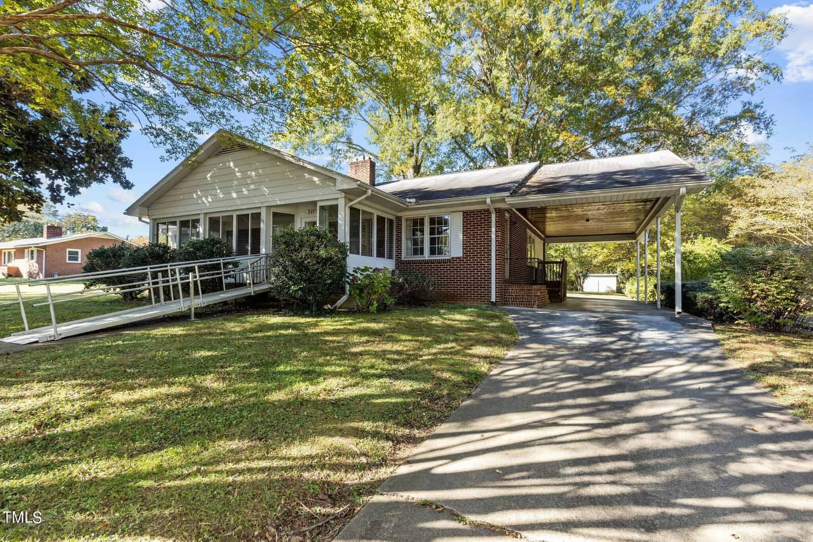 Nestled on 311 Delacroix Street, this Oxford brick house features a charming white porch and sloped ramp, embraced by lush green trees. A driveway guides you to the covered carport, and the home is perfectly set in a sunny, serene residential area.