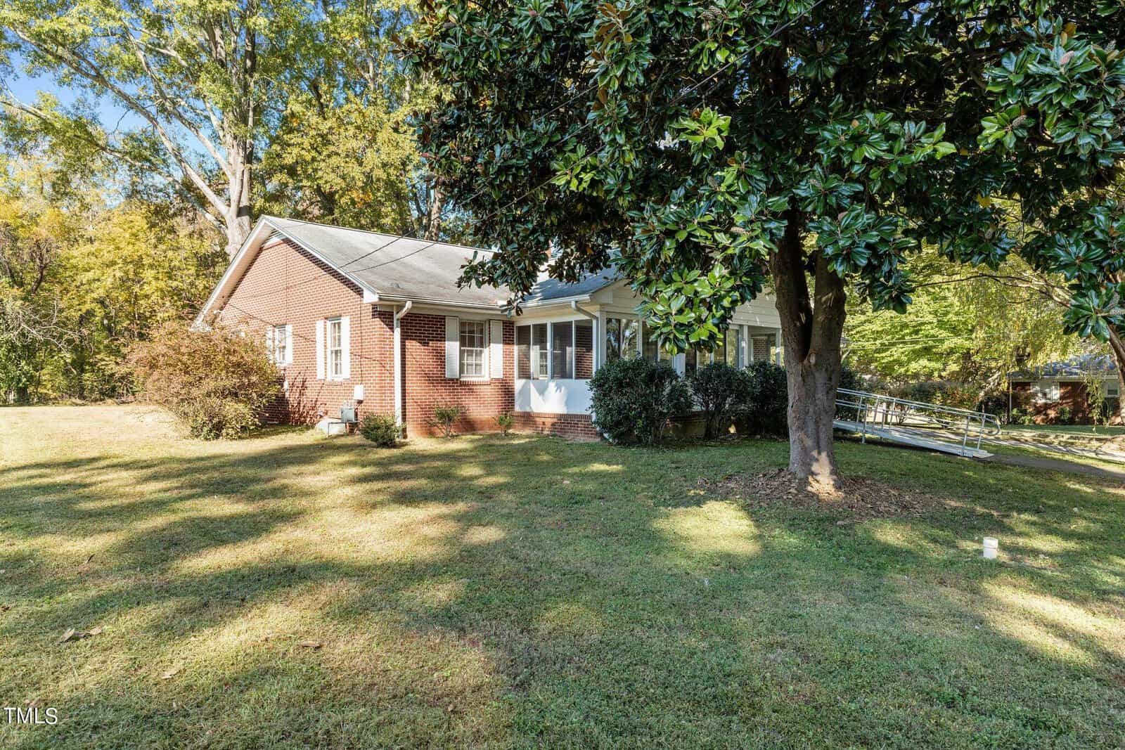 A brick house with a white roof sits amid lush green grass and surrounded by trees. It has a small front porch with steps leading up, and a ramp on one side. The sun casts shadows on the well-kept lawn.