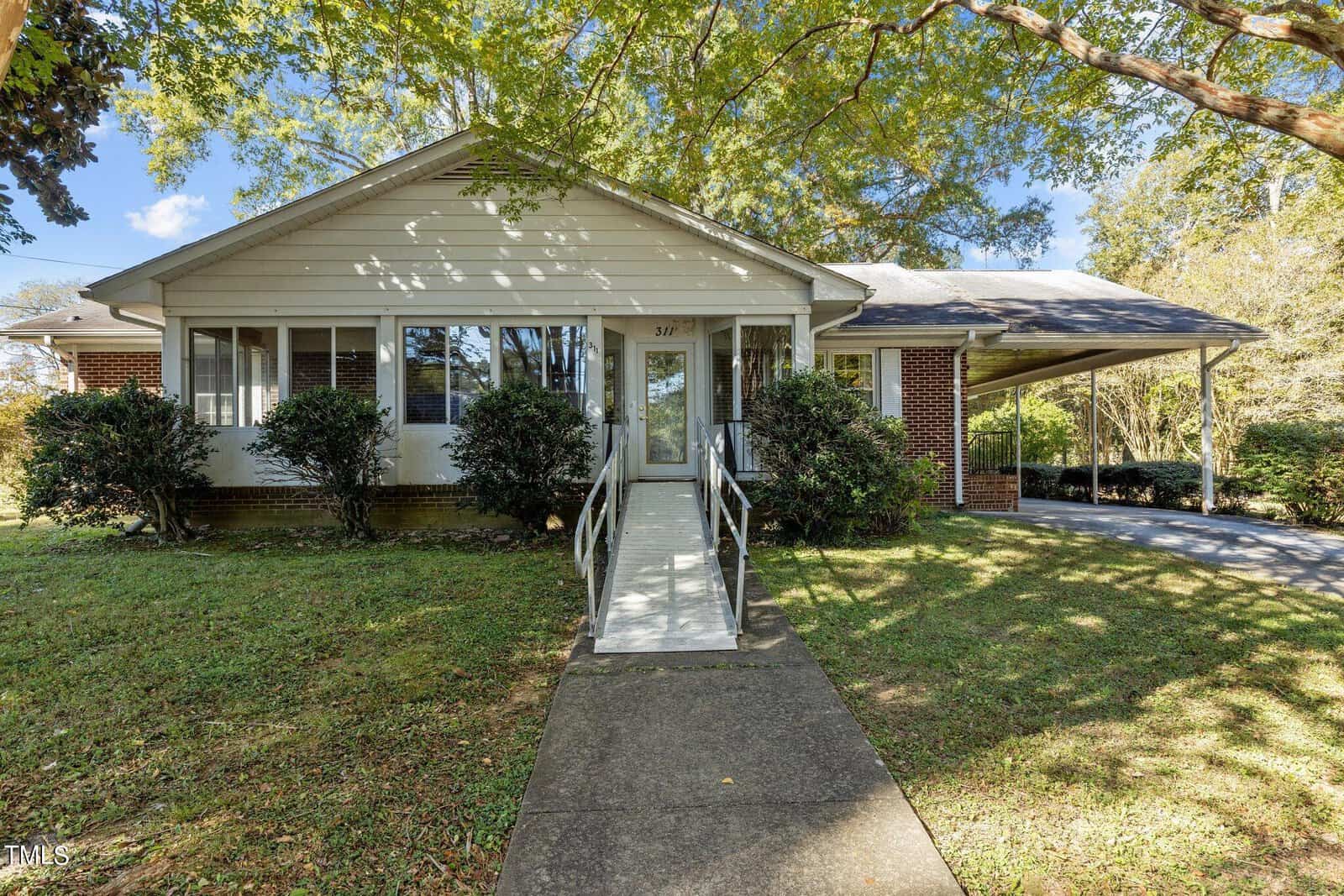 A single-story brick house with a white facade, fronted by lush greenery. A concrete path and ramp lead to the front entrance. Large trees provide shade, and a carport is visible on the right side.