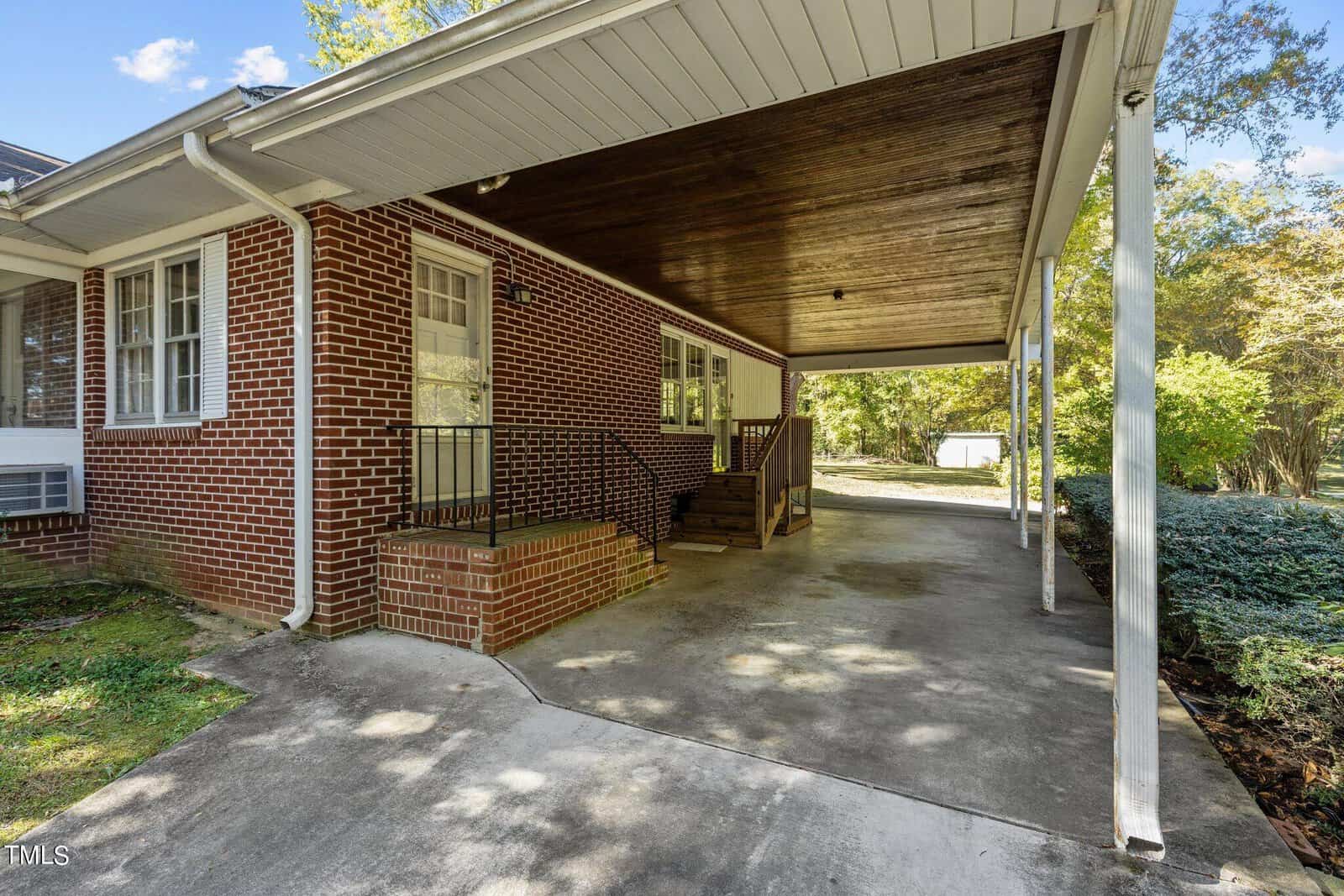 A brick house with a covered carport. Steps lead to a side entrance with a small porch. The driveway extends toward the background, bordered by grass and trees. The sky is partly cloudy.