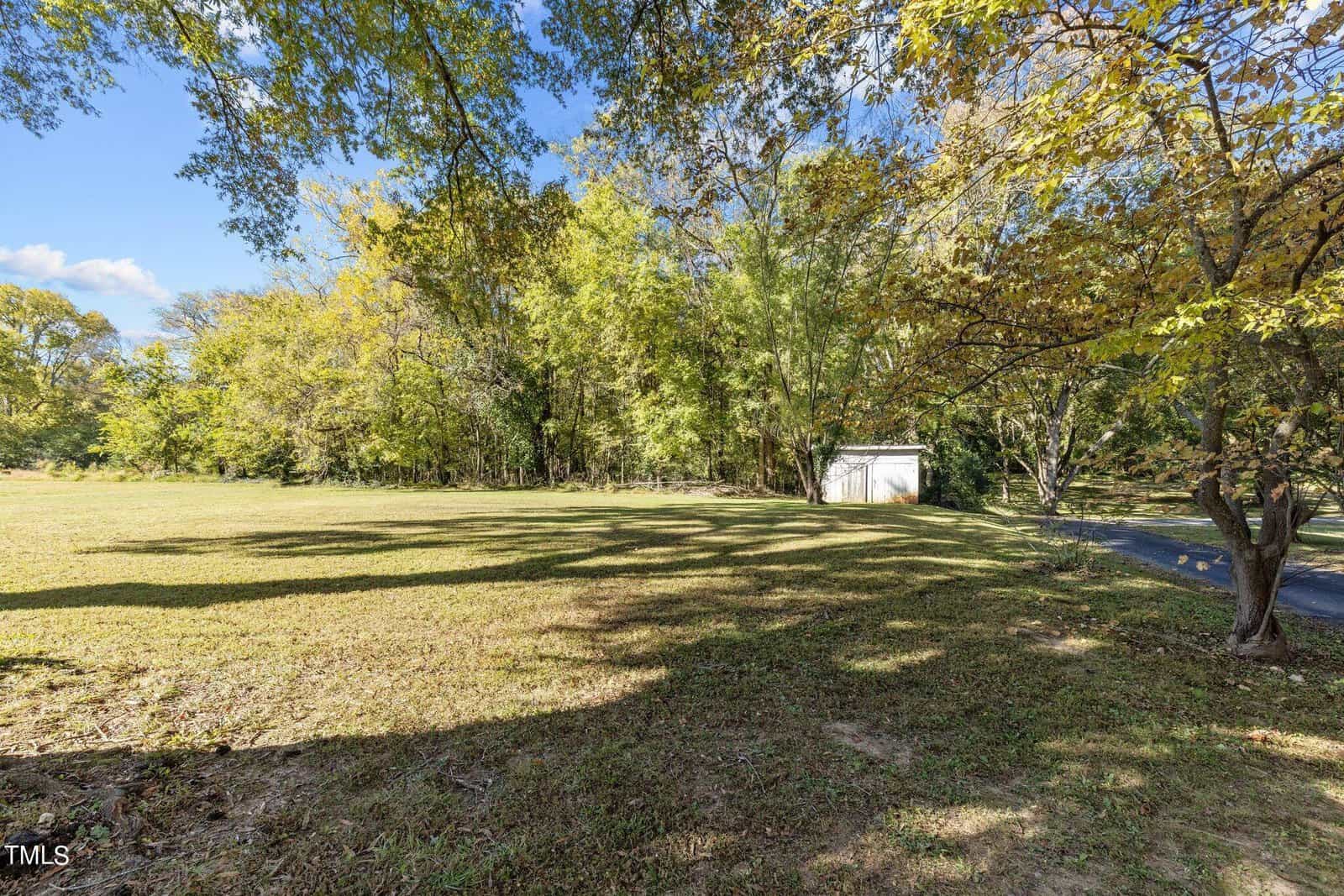 A grassy field bordered by trees with some autumn foliage, under a partly cloudy sky. A small white shed is visible near a paved road on the right side of the image.