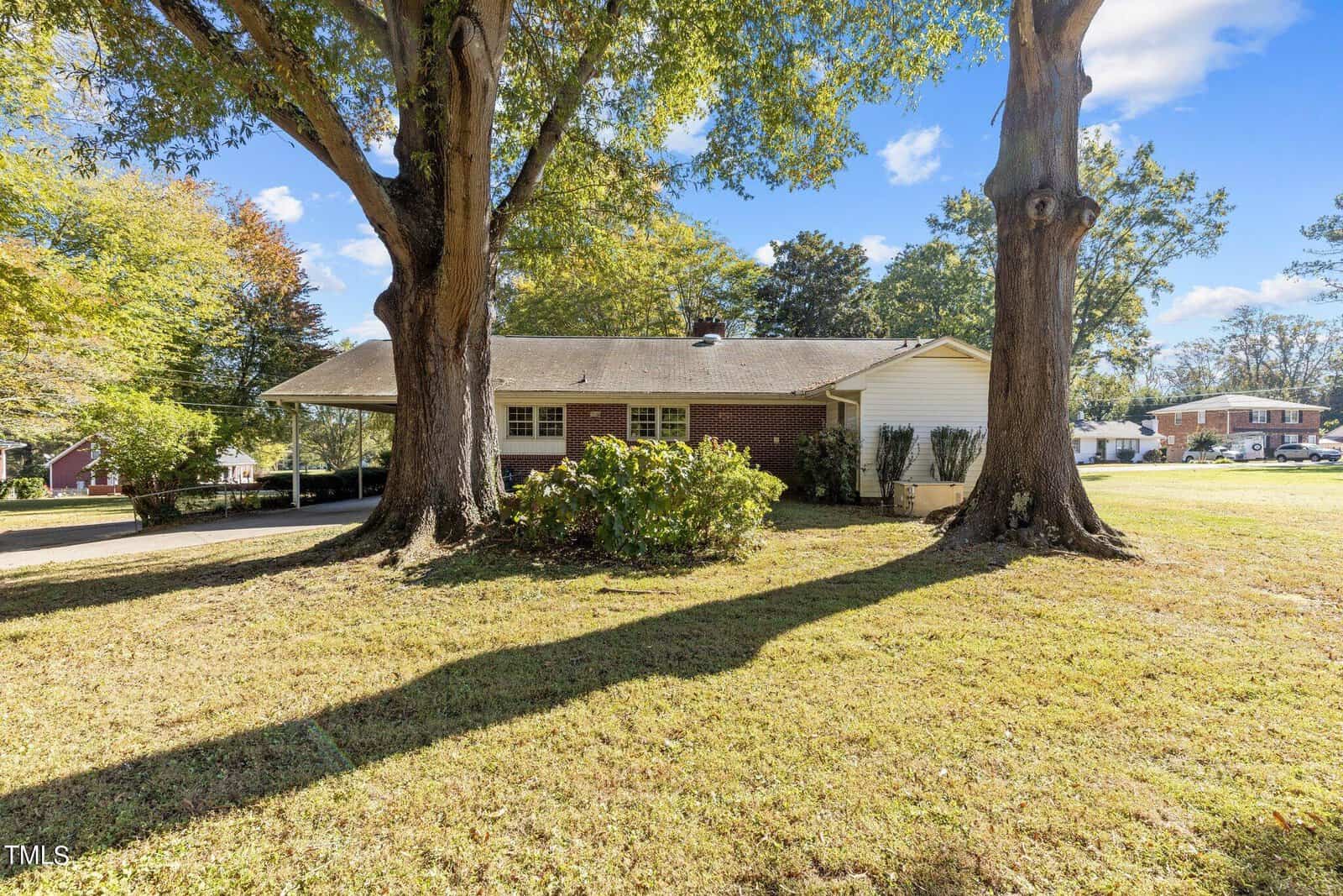 Front yard of a single-story brick house with two large trees in the foreground. A clear blue sky and a few clouds are visible in the background, with neighboring houses seen on the right side.