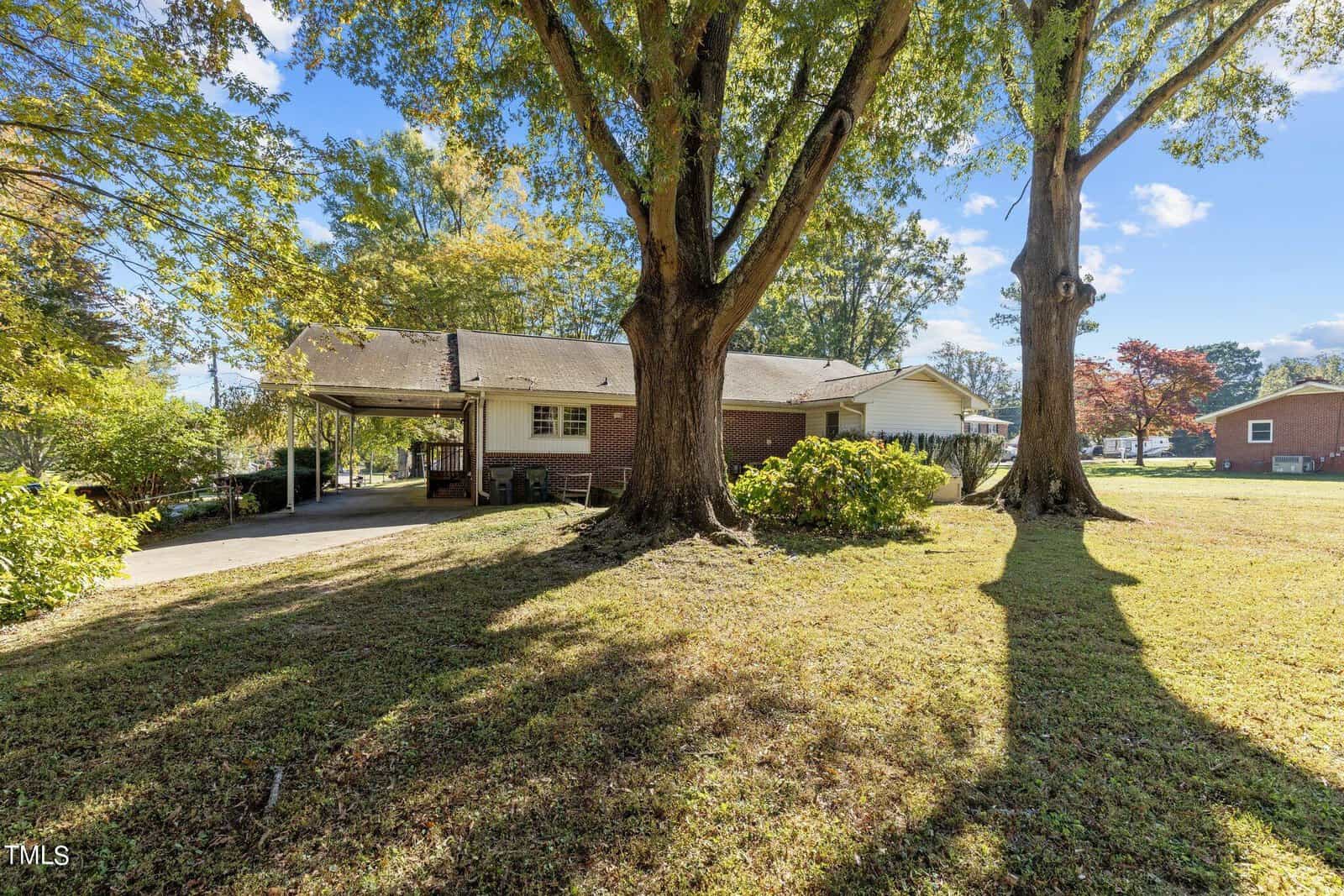 A scenic backyard with two large trees casting long shadows on the grassy lawn. The brick house in the background has a covered patio and is surrounded by lush greenery under a clear blue sky.