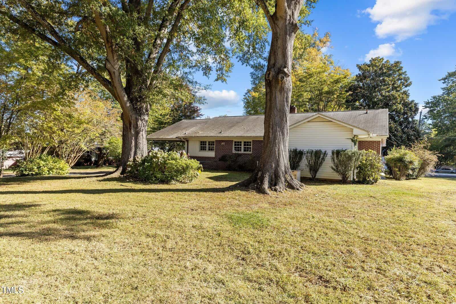 A single-story brick house with a gable roof is surrounded by large trees and a well-maintained lawn. The setting is sunny, with a clear blue sky and lush greenery, suggesting a serene suburban environment.