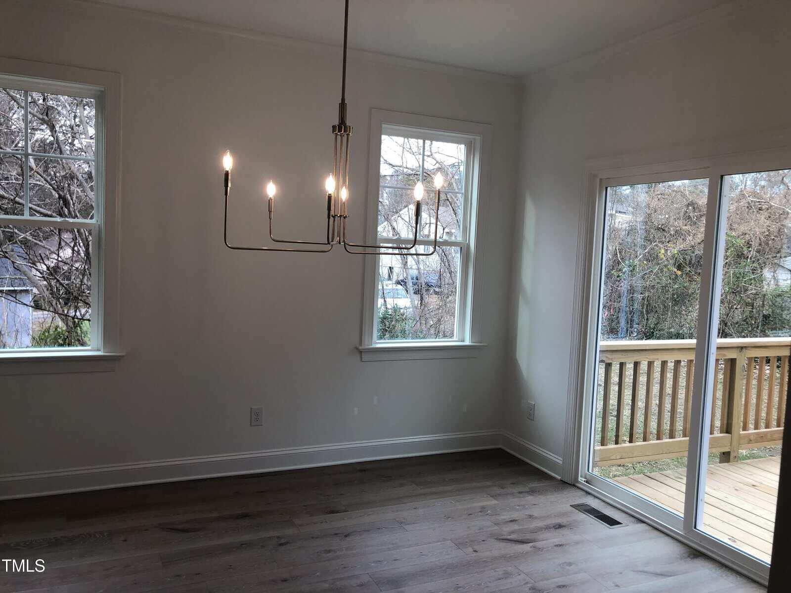 A modern, empty room with two large windows and a sliding glass door leading to a wooden deck. The room features a minimalist chandelier and hardwood floors. Bare trees are visible outside.