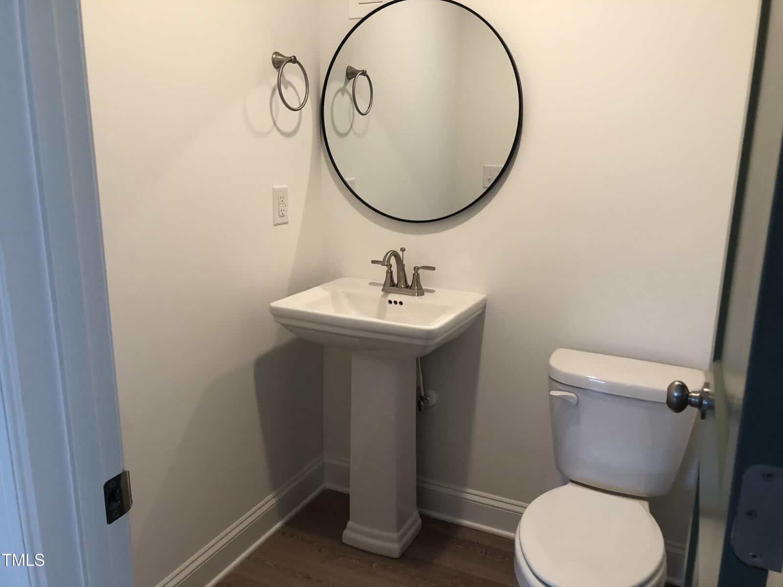 A small bathroom with a pedestal sink and a round mirror above it. There is a white toilet to the right and towel rings on the wall. The walls are light-colored, and the floor is wooden.
