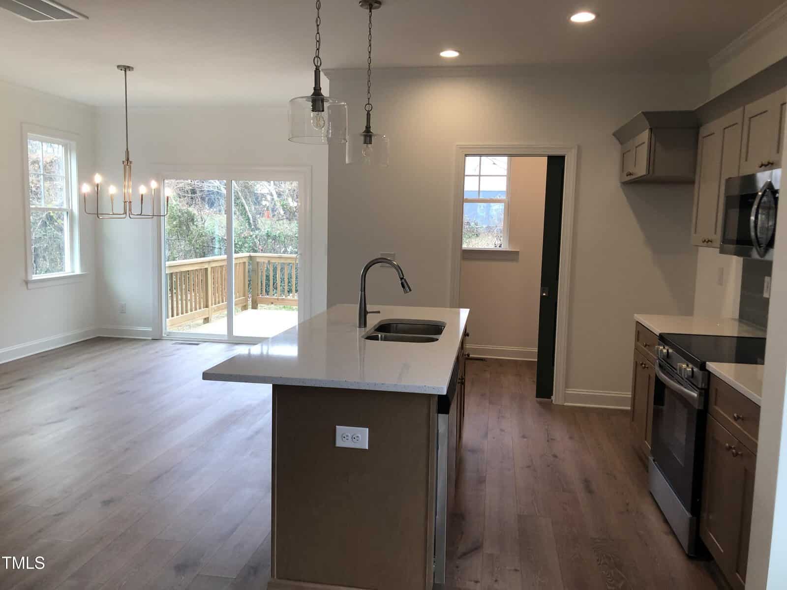 A modern kitchen with a central island and sink, pendant lights, and stainless steel appliances. Wood flooring extends to a dining area with a chandelier and large windows. Glass doors open to a deck with a view of trees outside.