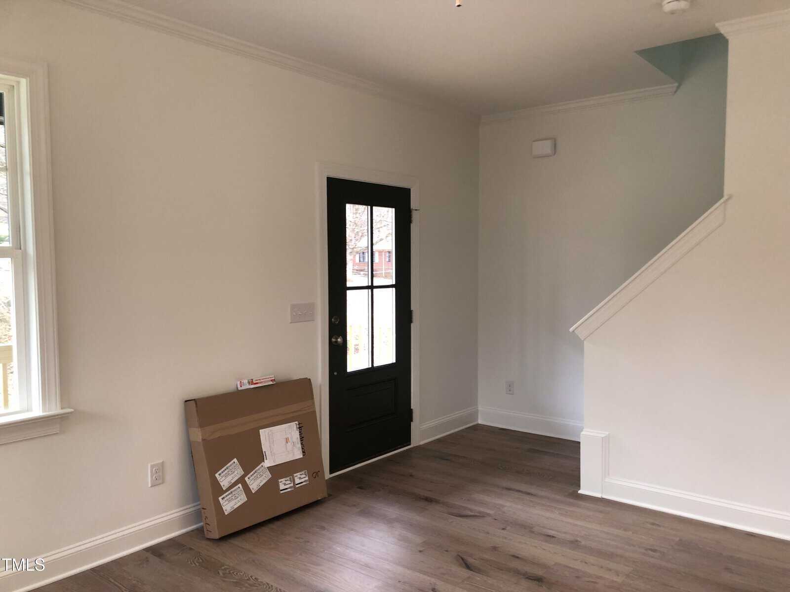 A minimalist room with light wood flooring and white walls features a black front door with glass panes. A cardboard box rests against the wall near the door. A window is on the left, and a staircase leads upwards on the right.