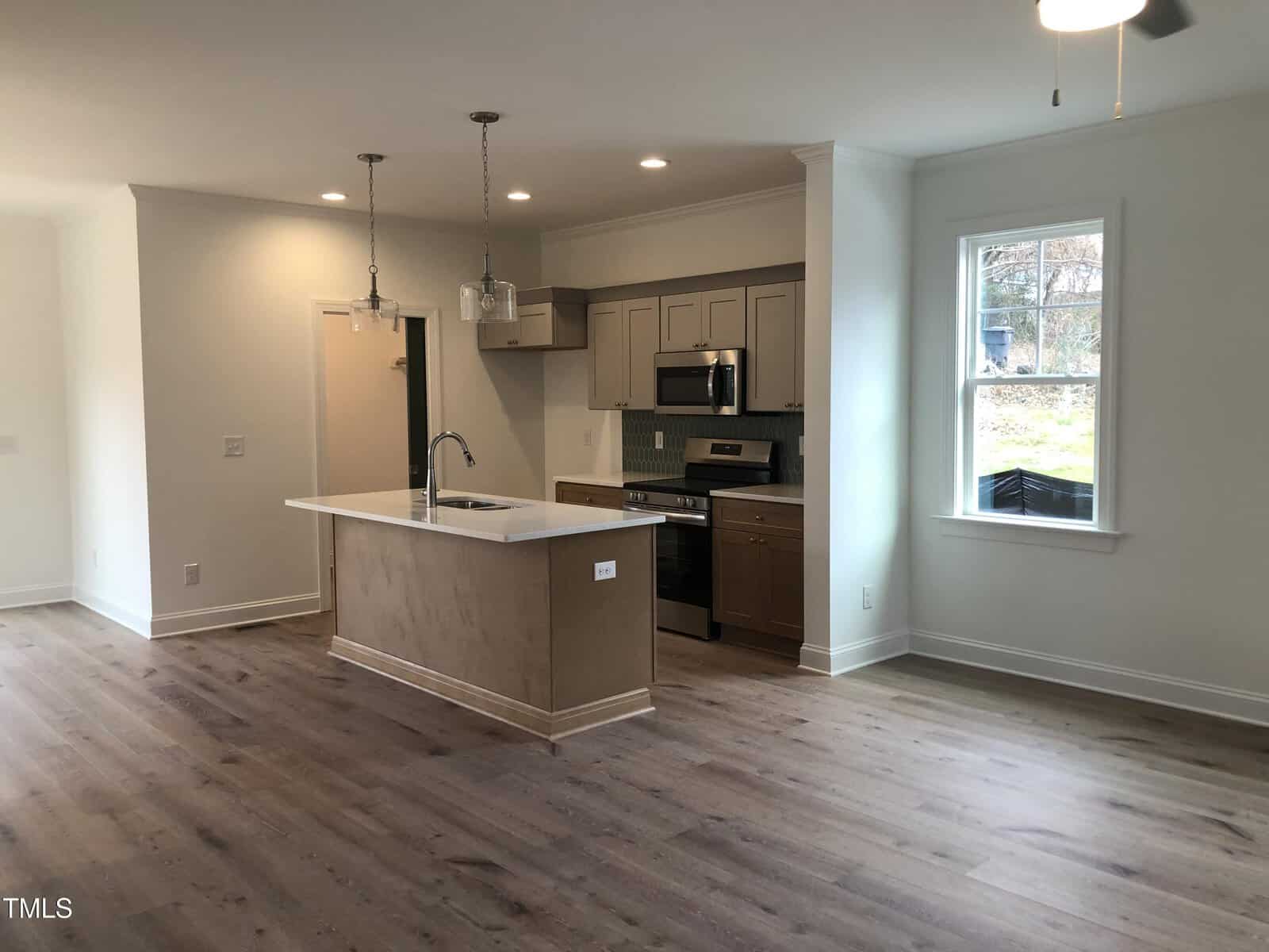 A modern kitchen with wooden flooring, featuring a central island with a sink and pendant lights overhead. The kitchen has gray cabinets, a stainless steel stove, and a range hood. A window allows natural light to illuminate the space.