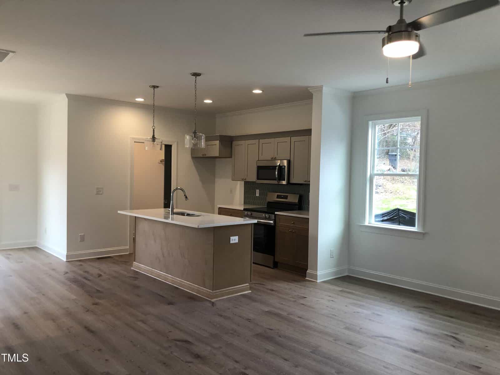 A modern, unfurnished kitchen and living area with light wood flooring. Features include an island with a sink, overhead pendant lights, stainless steel appliances, and a ceiling fan. A window on the right brings in natural light.