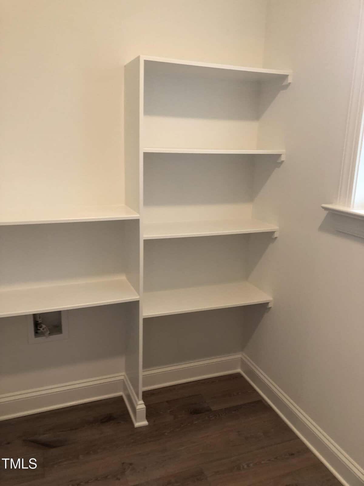 Empty white built-in shelves against a light-colored wall, with a small square space possibly for an appliance, on a room corner with wooden flooring.