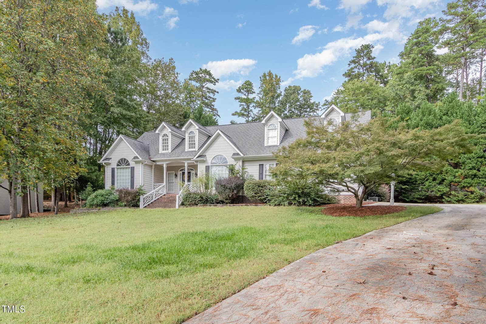A large white house with a gabled roof is surrounded by trees and a well-kept lawn. A curved driveway leads to the entrance, and there's a mix of greenery and shrubs in the front yard. The sky is partly cloudy, with a few patches of blue.