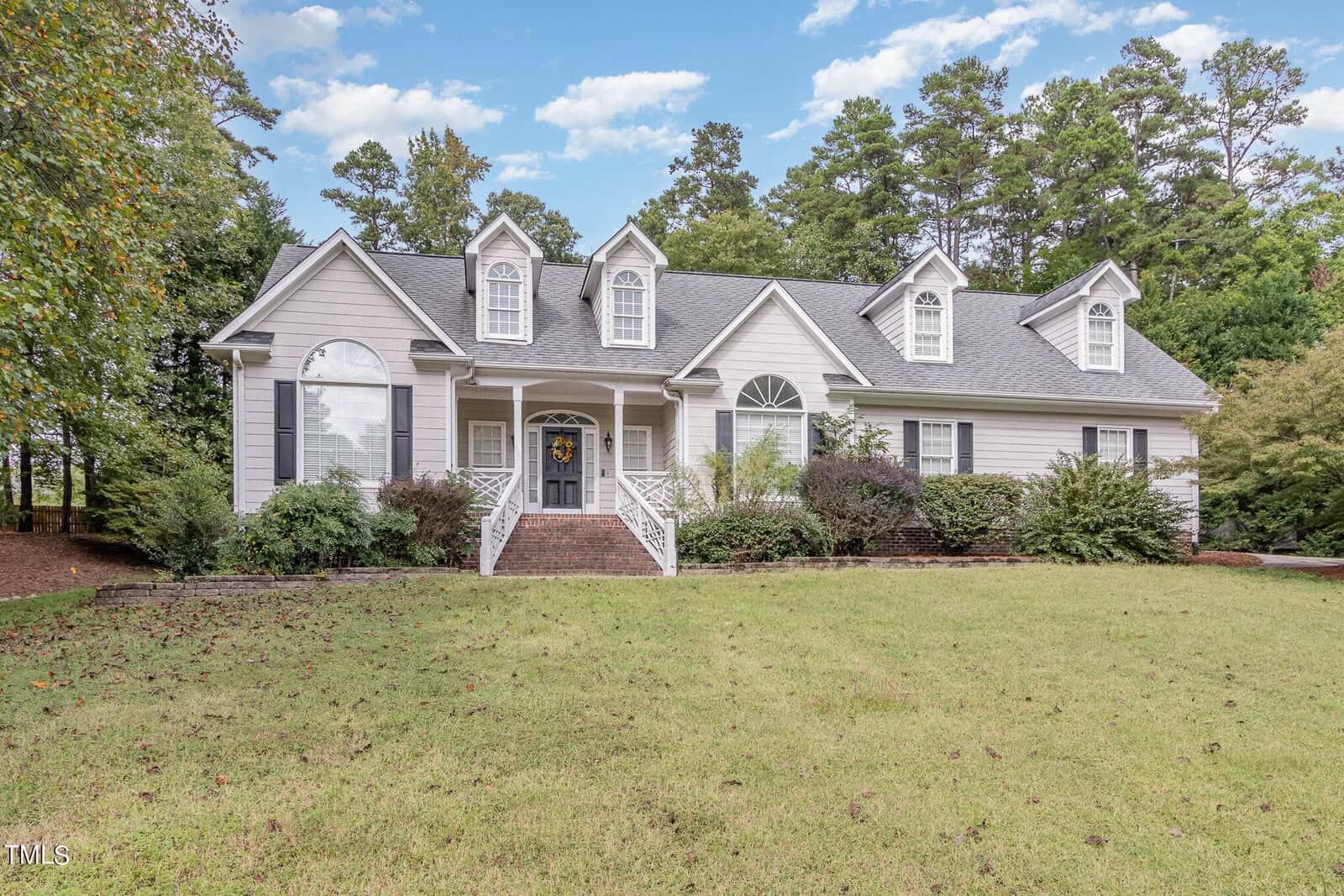 A two-story suburban house at 1102 Closs Court boasts gray siding and white trim, featuring three dormer windows on the roof. A brick walkway leads to the entrance with a wreath on the door. Surrounded by a lawn and trees under Henderson's partly cloudy sky, it exudes charm.