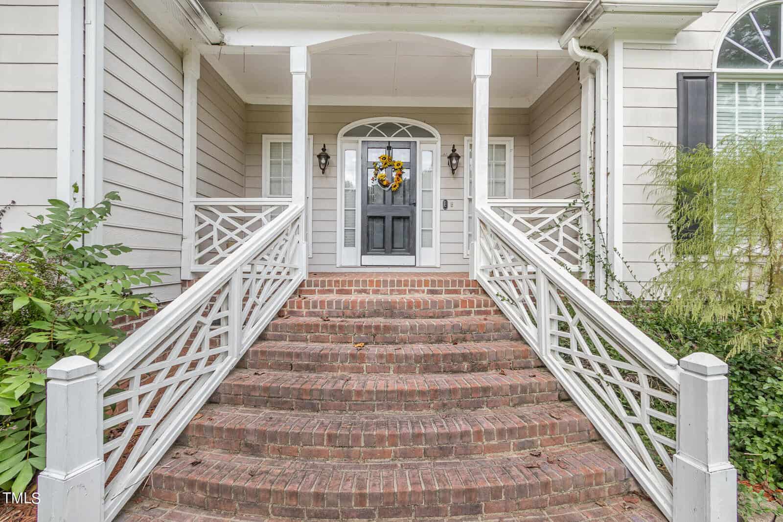 Front entrance of a house with a welcoming black door adorned with a bright floral wreath. Brick steps lead up to the porch, bordered by white railings. The exterior is light grey with decorative trim. Lush greenery flanks the steps.
