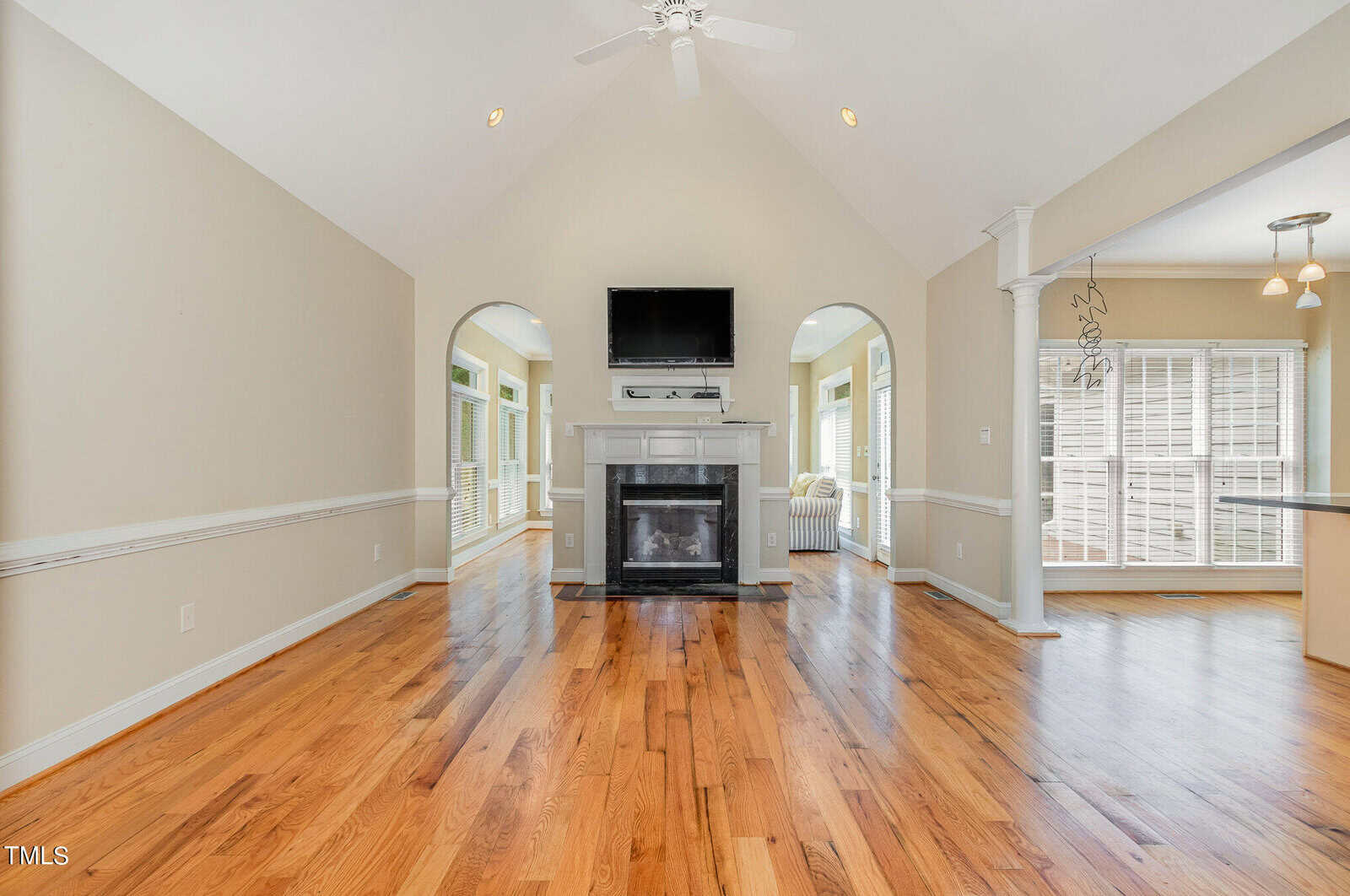 Bright, spacious living room with vaulted ceiling features a ceiling fan and a fireplace centered between two arched doorways. Hardwood floors extend to a sunlit seating area with large windows. A glimpse of the adjacent kitchen is visible.