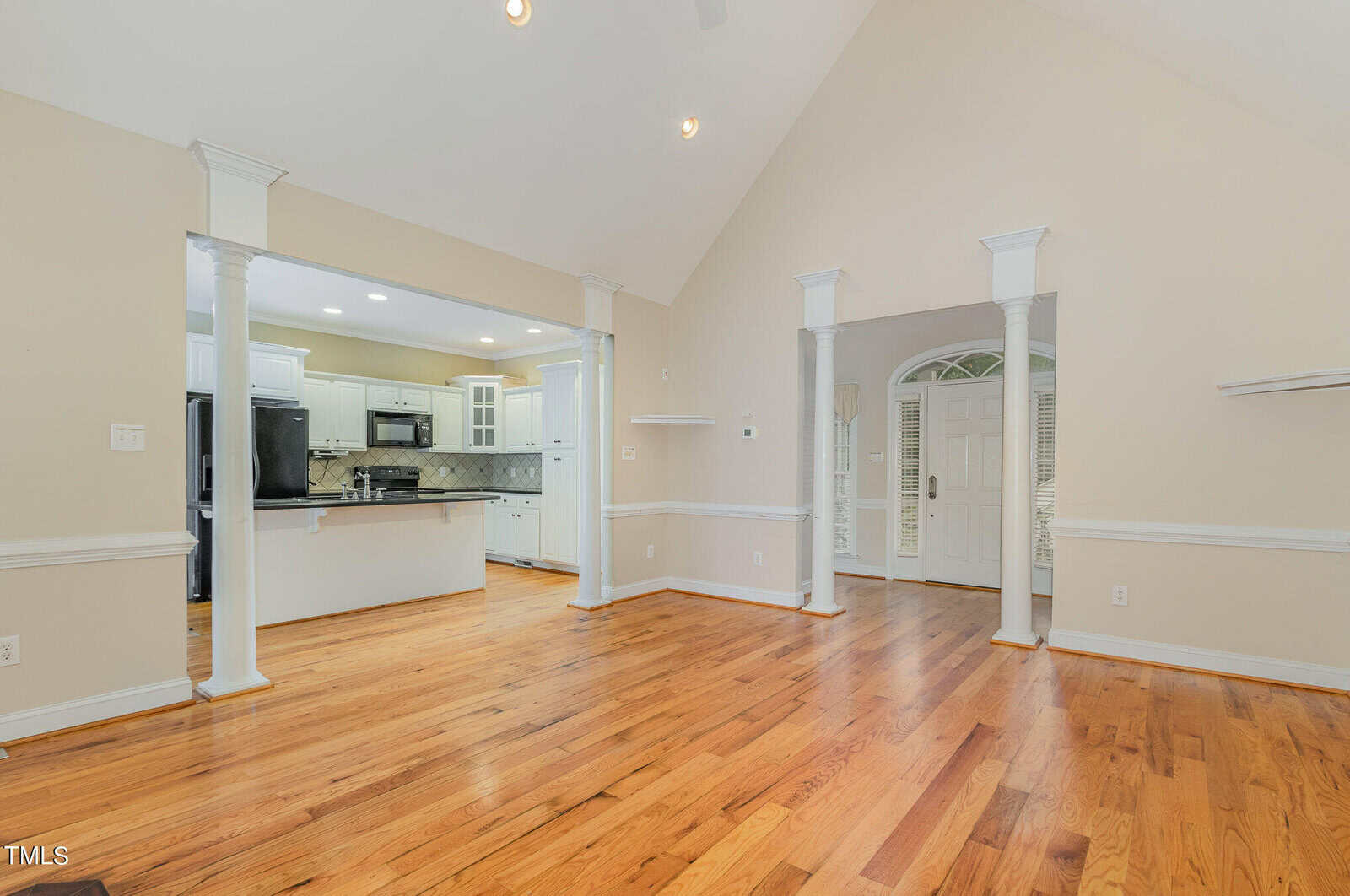 Spacious room with polished hardwood floors and a vaulted ceiling. Open layout leads to a modern kitchen with white cabinetry and stainless steel appliances. Entryway with a decorative glass-panel door and sidelights. White pillars and trim accent the space.