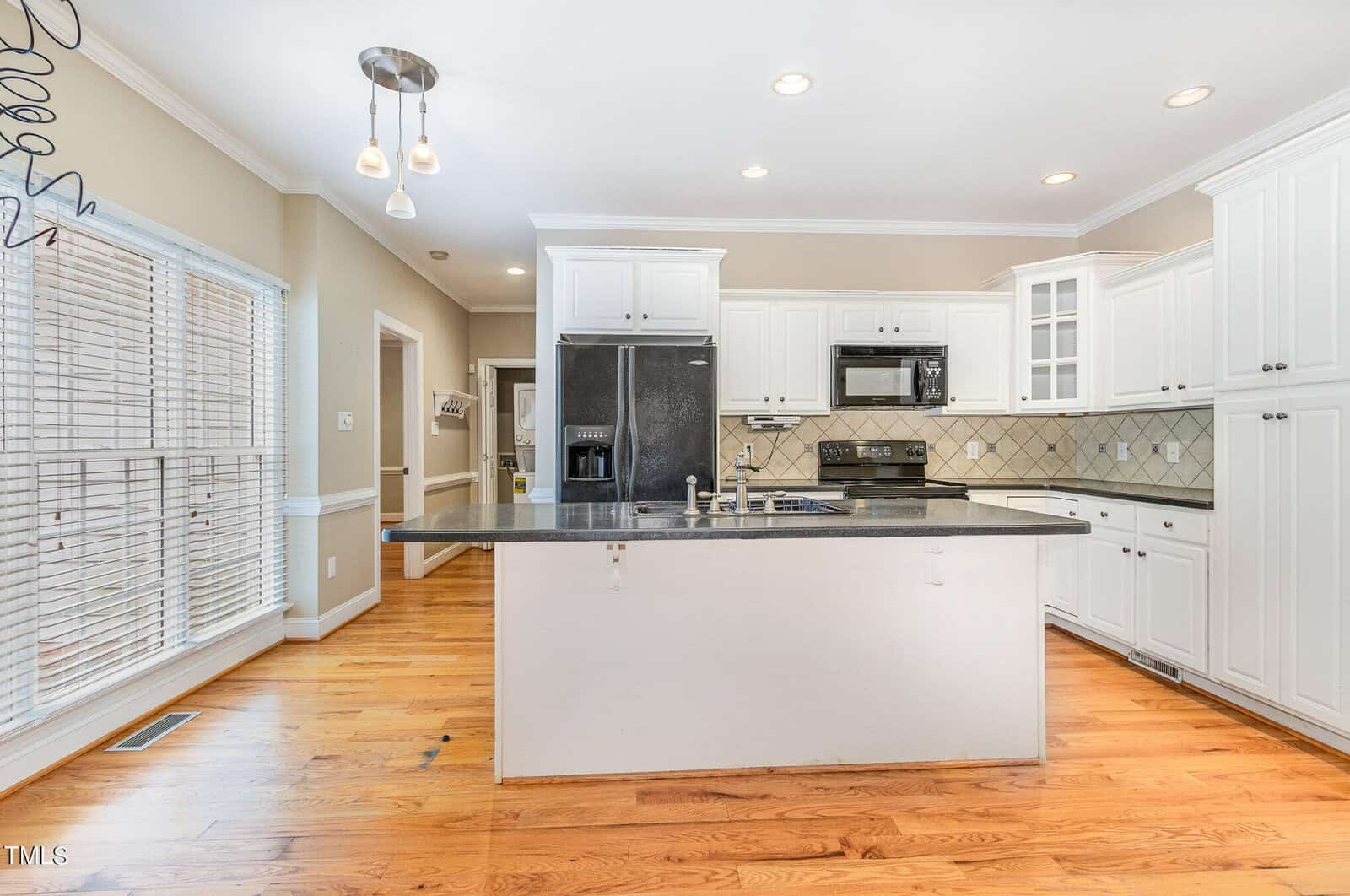 A bright kitchen with white cabinets, a black countertop island, and stainless steel appliances. Hardwood floors add warmth, while large windows allow natural light to fill the space. Pendant lights hang above the island.