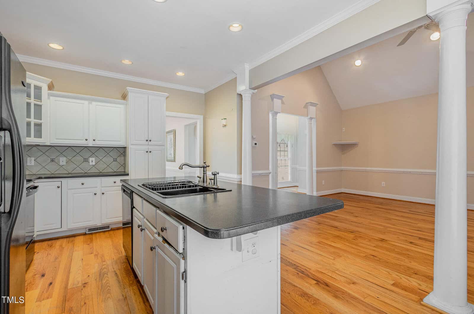 A spacious kitchen with white cabinets and a black countertop island. The room features hardwood floors and recessed lighting. Adjacent is a dining area with high ceilings and two white columns, leading to another bright, open space.