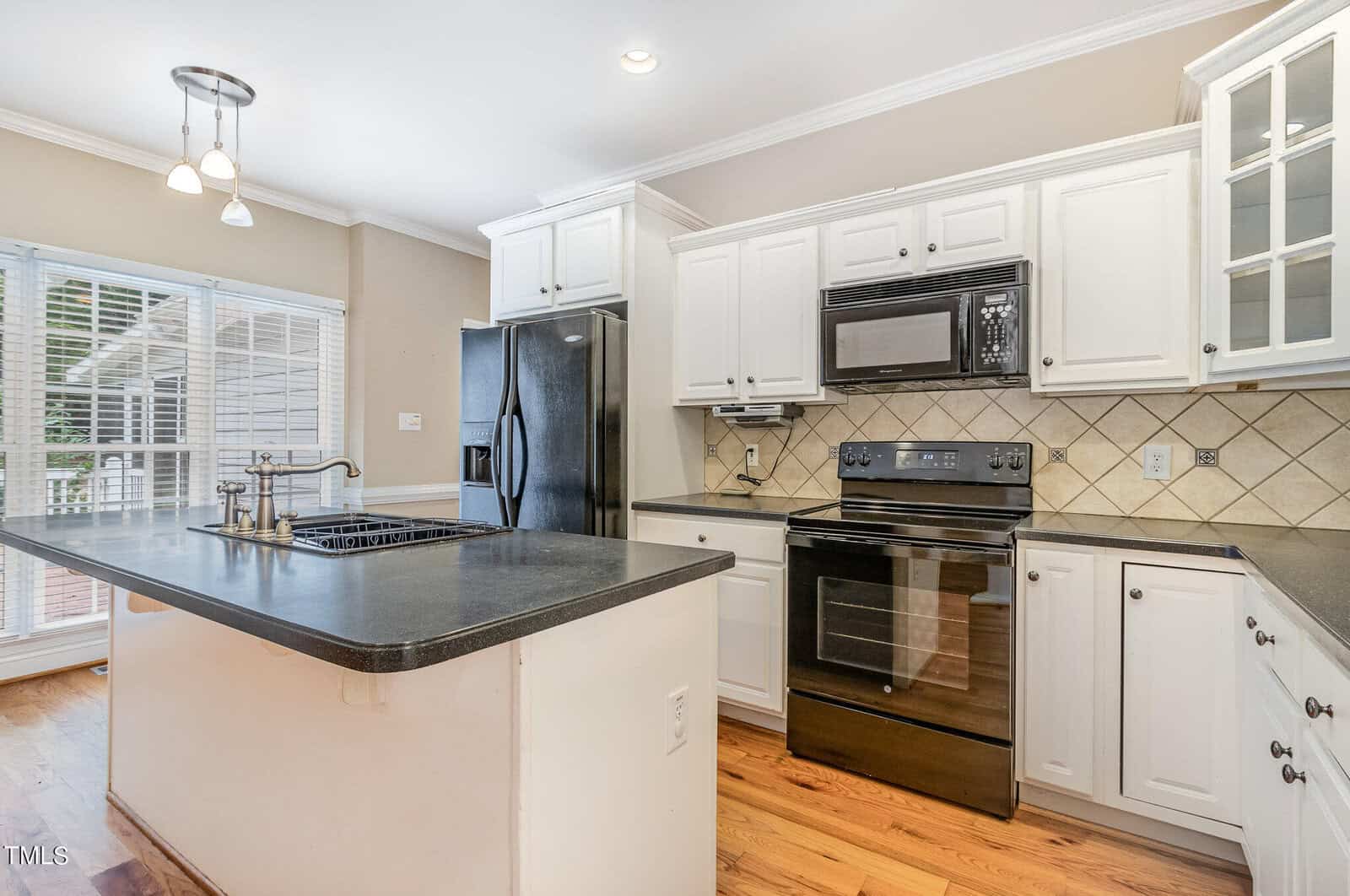 A modern kitchen with white cabinets, a black countertop, and a black stove and refrigerator. There's a kitchen island with a sink and light fixtures above. Hardwood floors and large windows bring in natural light.