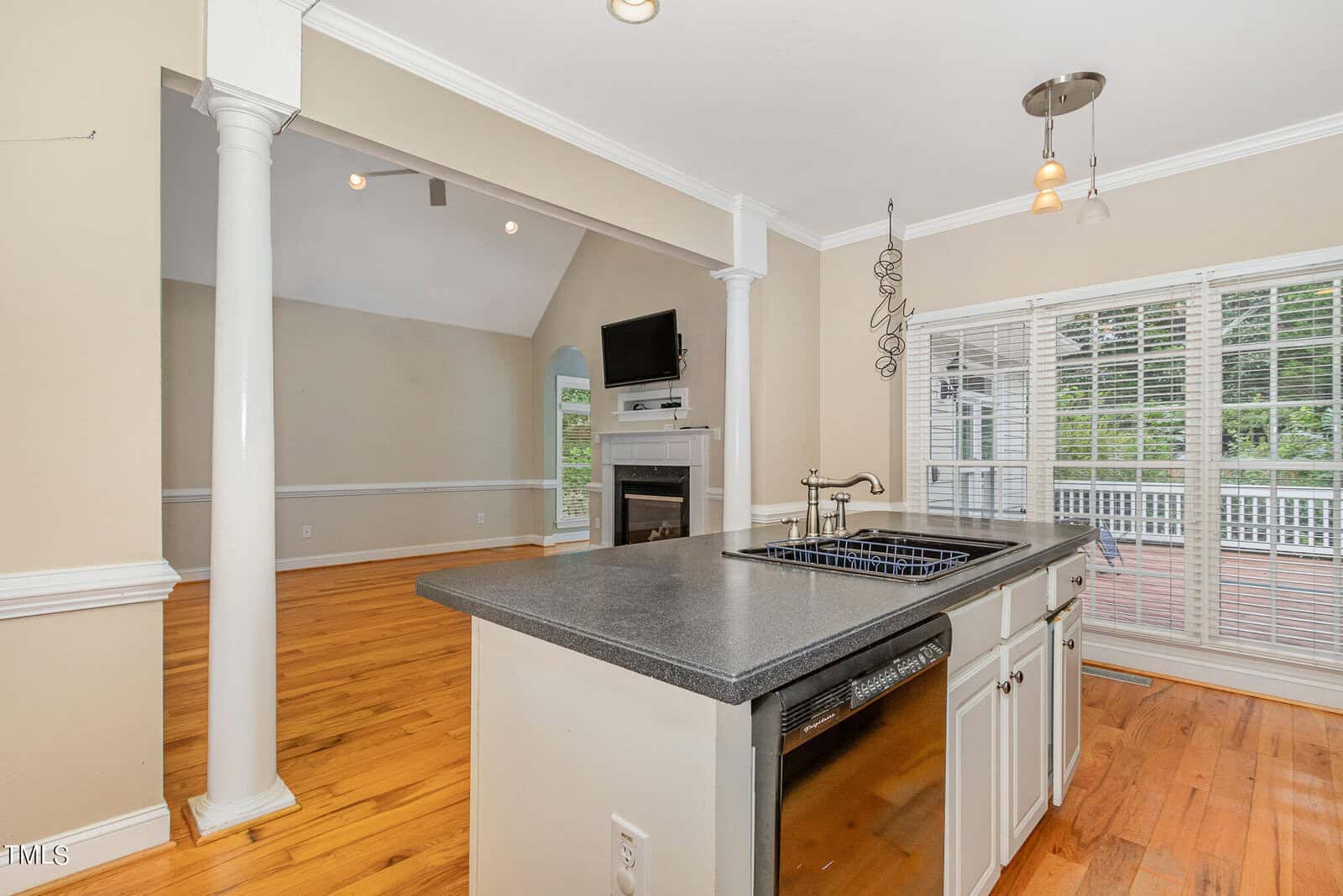 Bright and spacious kitchen and living area with hardwood floors, a large kitchen island with a sink, and white cabinetry. The room features pillars, a fireplace, a wall-mounted TV, and large windows with views of a deck and greenery outside.