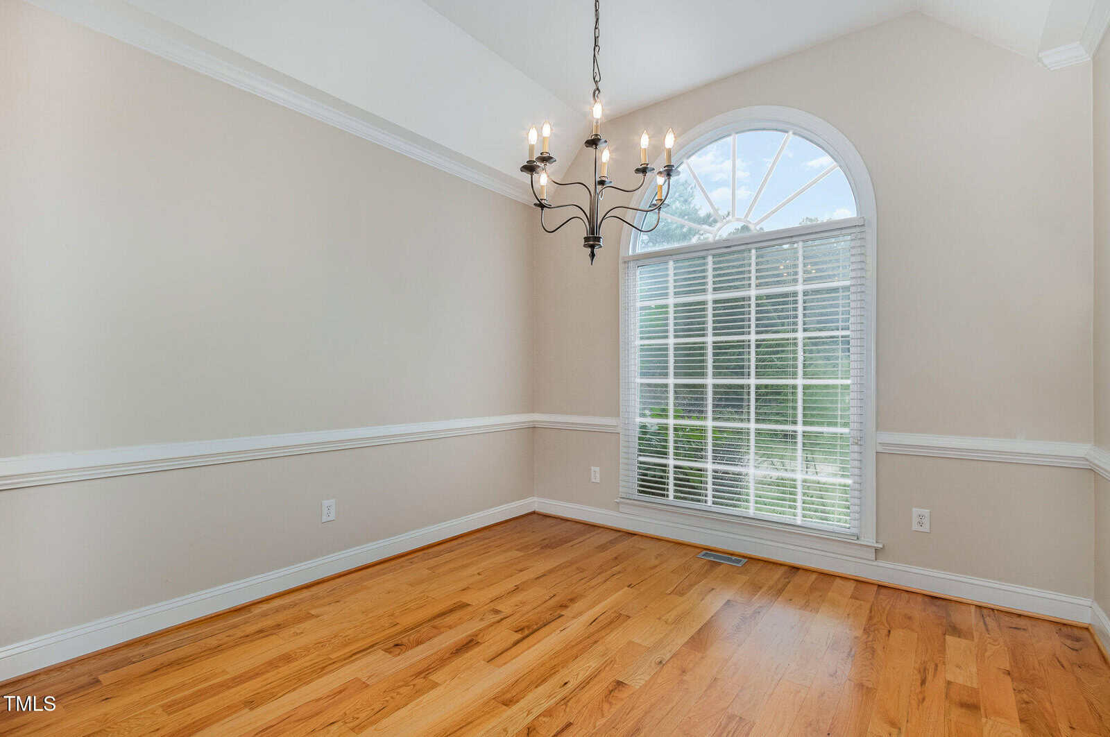 An empty room with light-colored walls and a wooden floor. It features a large arched window with blinds, allowing natural light to illuminate the space. A black, five-arm chandelier hangs from the ceiling.