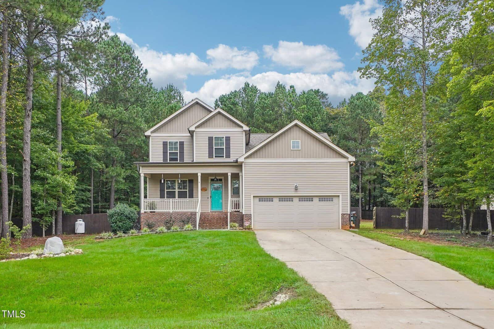 Located at 2129 Emerald Lane in the heart of Franklinton, this two-story house boasts a beige exterior and a vibrant teal front door, nestled on a lush green lawn. The driveway guides you to a spacious double garage, all framed by majestic trees and a partly cloudy sky.