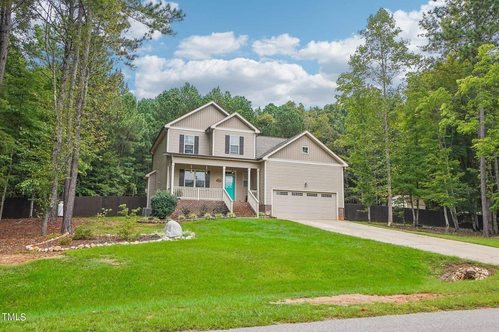 Two-story beige house with a turquoise front door, attached two-car garage, and neat lawn. The house is surrounded by trees and has a stone pathway leading to the entrance. A long driveway extends from the road to the garage.