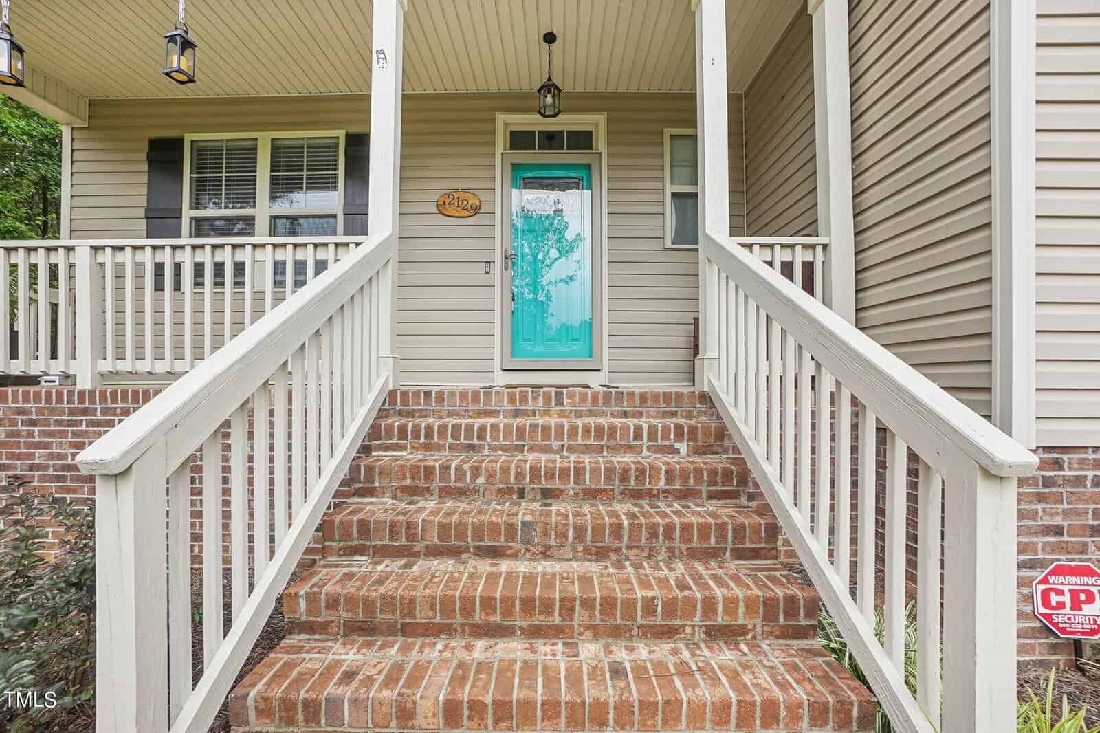 Staircase leading to a front porch with a turquoise door. The house exterior is beige with white railings and a brick foundation. A decorative hanging light and a small security sign are visible.