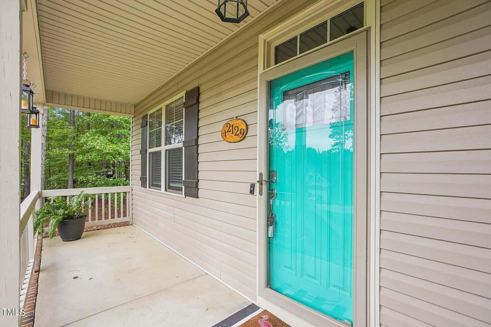 A house porch with a teal-colored front door, marked with the number 429. The porch has a covered roof, beige siding, a lantern-style light fixture, and a window with blinds. A potted plant is placed beside the railing. Trees are visible in the background.