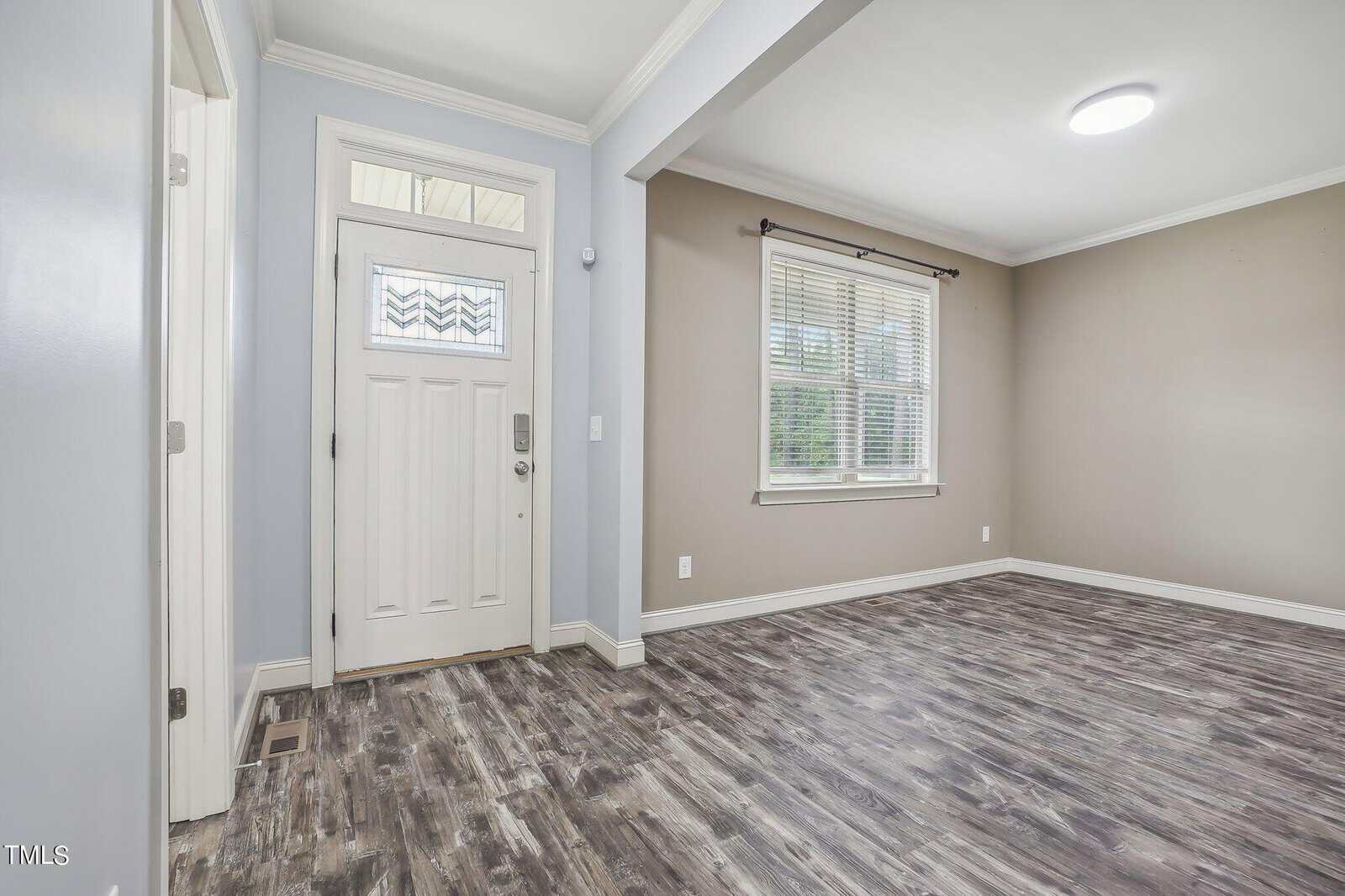 An empty living room with light gray walls and wood-patterned vinyl flooring. A single large window with blinds is on the right wall. The entrance door, painted white, is to the left, under recessed lighting.