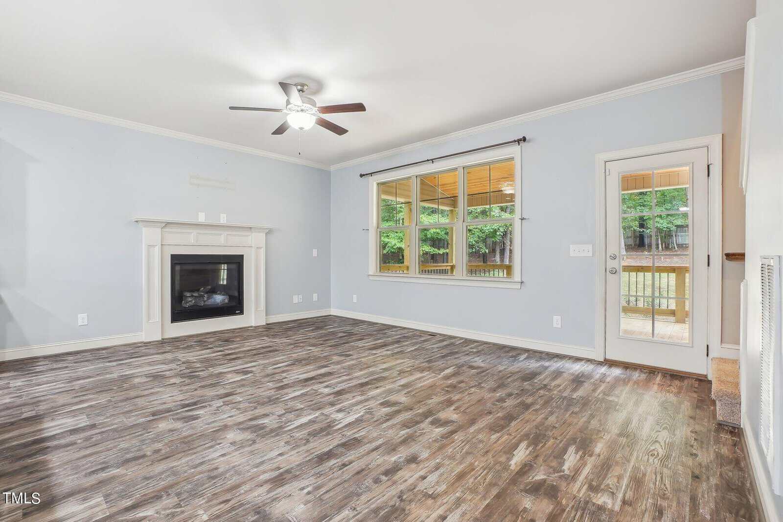 A spacious living room with light blue walls, a ceiling fan, and a fireplace on the left. Large window and door on the right, leading to a wooden deck. The floor is dark wood, and the room is well-lit with natural light.