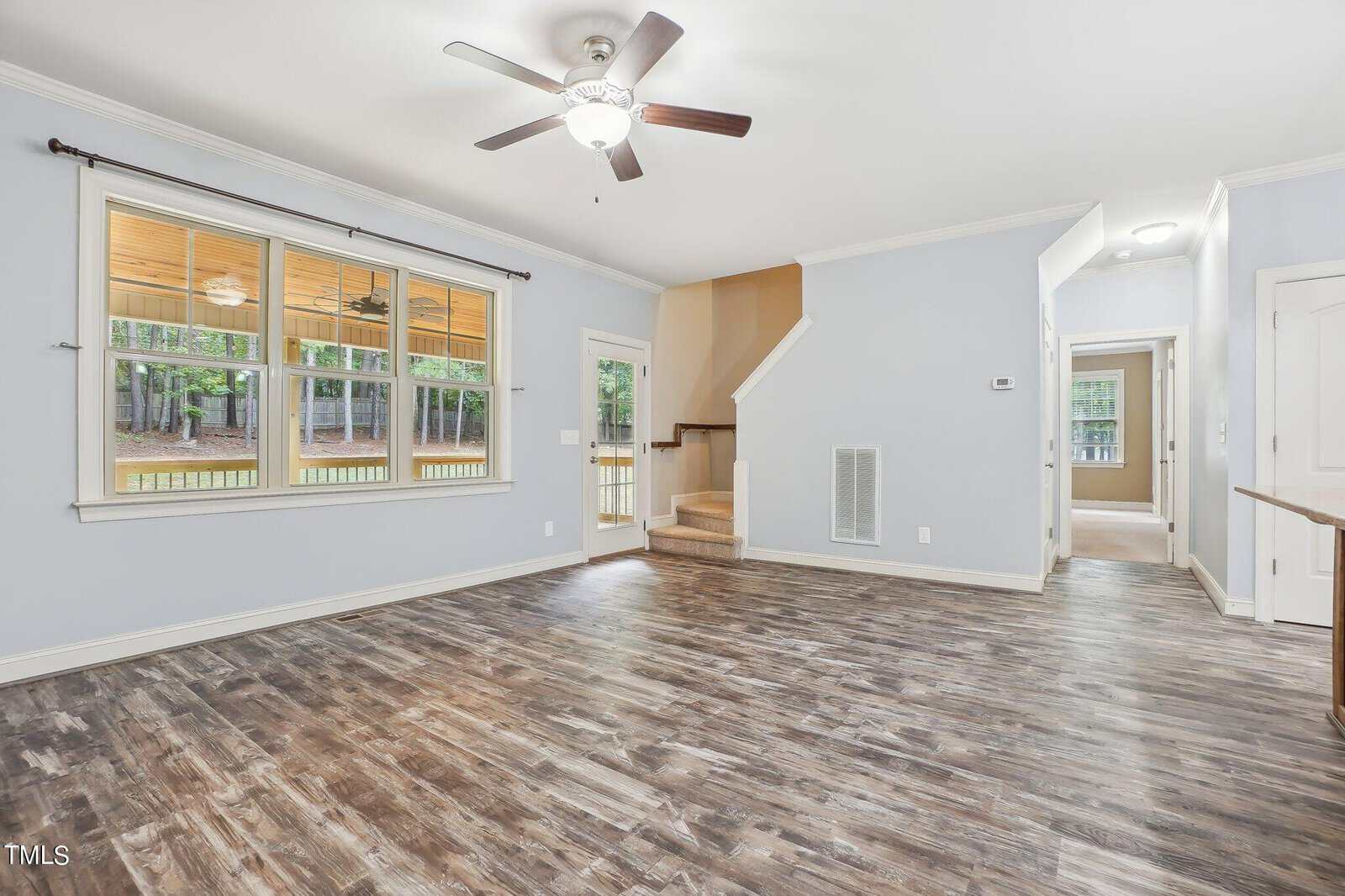 A spacious, empty living room with light blue walls and wood-look flooring. Features include a ceiling fan, large window with a view of a porch and trees, and a staircase leading upstairs. A hallway and a door to another room are visible.