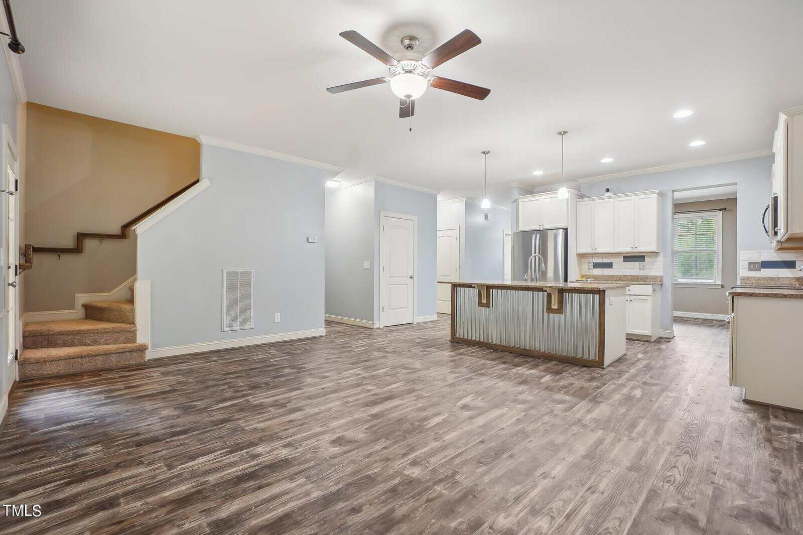 Spacious open-concept living area with wood flooring, ceiling fan, modern kitchen island, stainless steel appliances, white cabinetry, and recessed lighting. A staircase leads to the upper level. Natural light enters through a window in the kitchen.