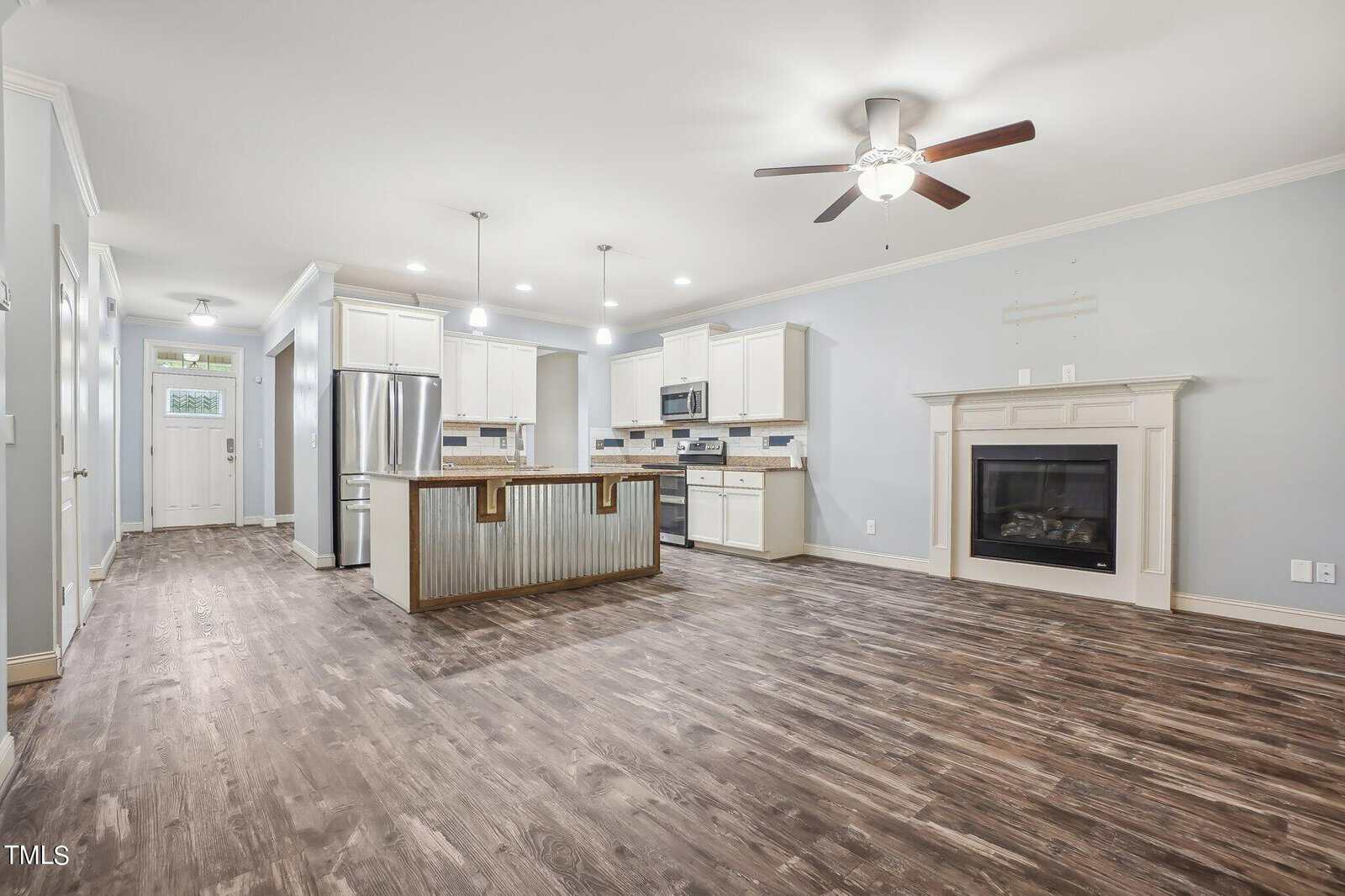 A spacious, empty kitchen and living area with wood flooring. The kitchen features white cabinets, stainless steel appliances, and an island with a wooden accent. The living area has a ceiling fan and a fireplace with a mantle.