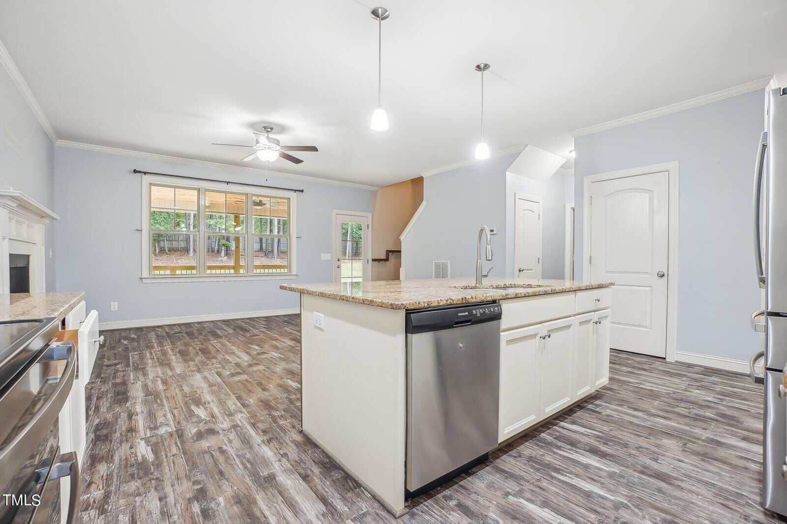 A modern kitchen and living area with wooden flooring, light blue walls, and white cabinets. It features a central island with a dishwasher, stainless steel appliances, and a ceiling fan. Large windows provide natural light. Stairs are visible in the background.