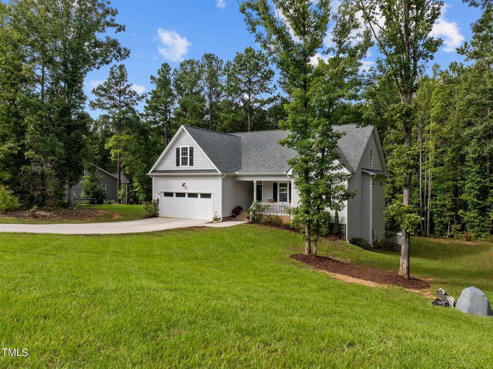 A modern white house with gray roofing, featuring a two-car garage. It's surrounded by a lush green lawn and tall trees. The sky is clear with a few clouds, creating a serene, suburban setting.