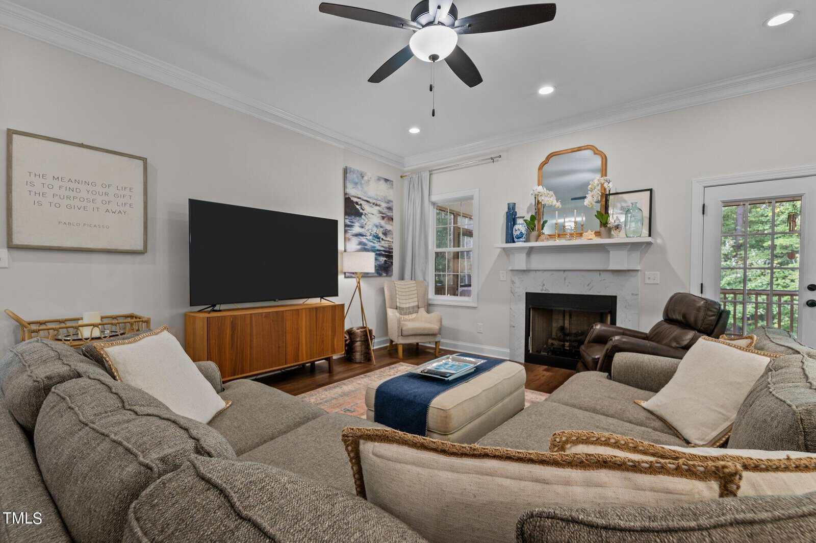 A cozy living room with a gray sectional sofa, a wooden TV stand with a large flat-screen TV, framed art on the walls, a fireplace with decor, an armchair, and a ceiling fan. A door leads outside, with greenery visible through the windows.