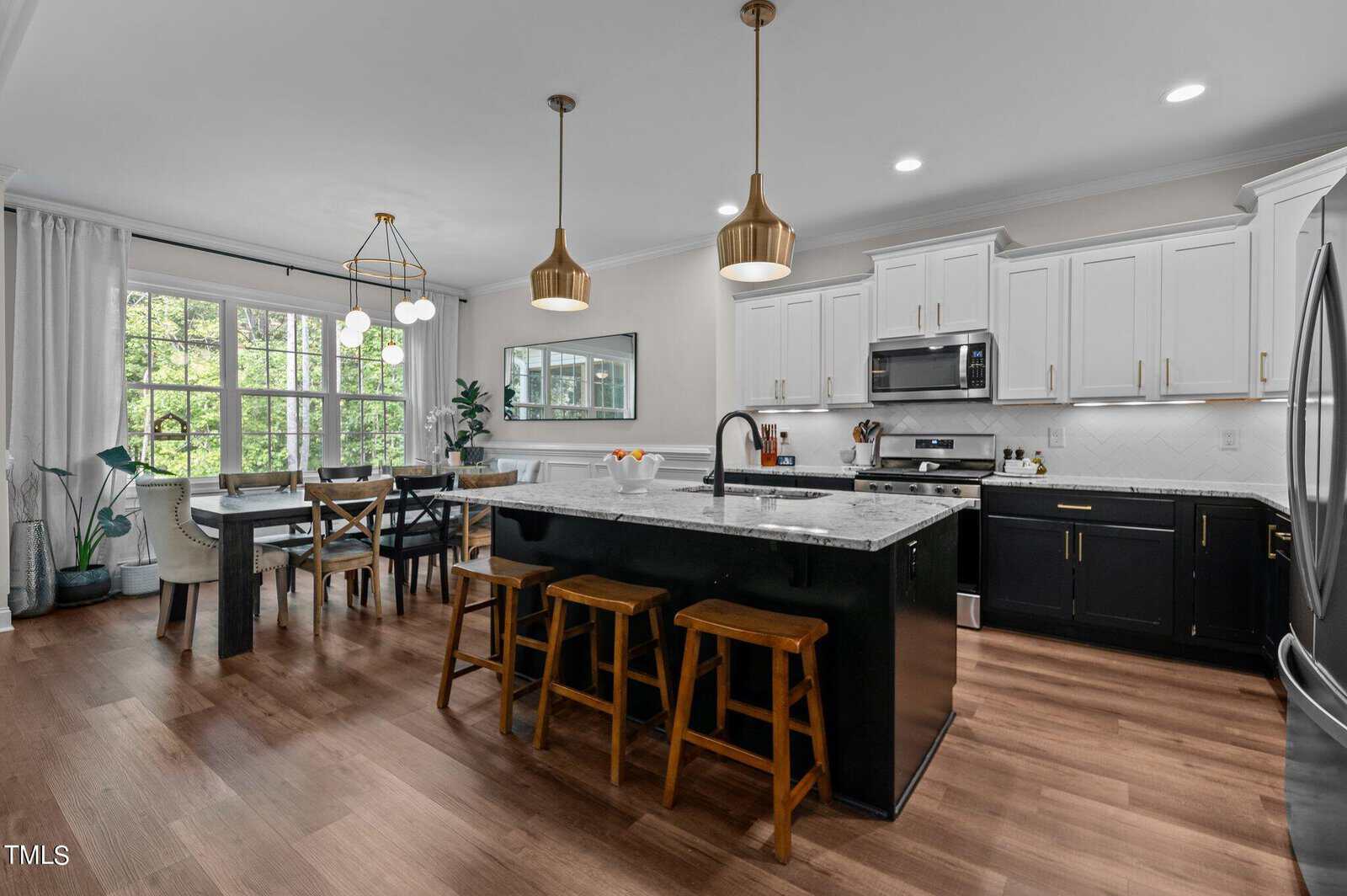 Modern kitchen and dining area with a large island, marble countertops, and wooden bar stools. White cabinets contrast with dark lower cabinets. Gold pendant lights hang above, and a dining table by the window has upholstered chairs. Greenery accents the space.