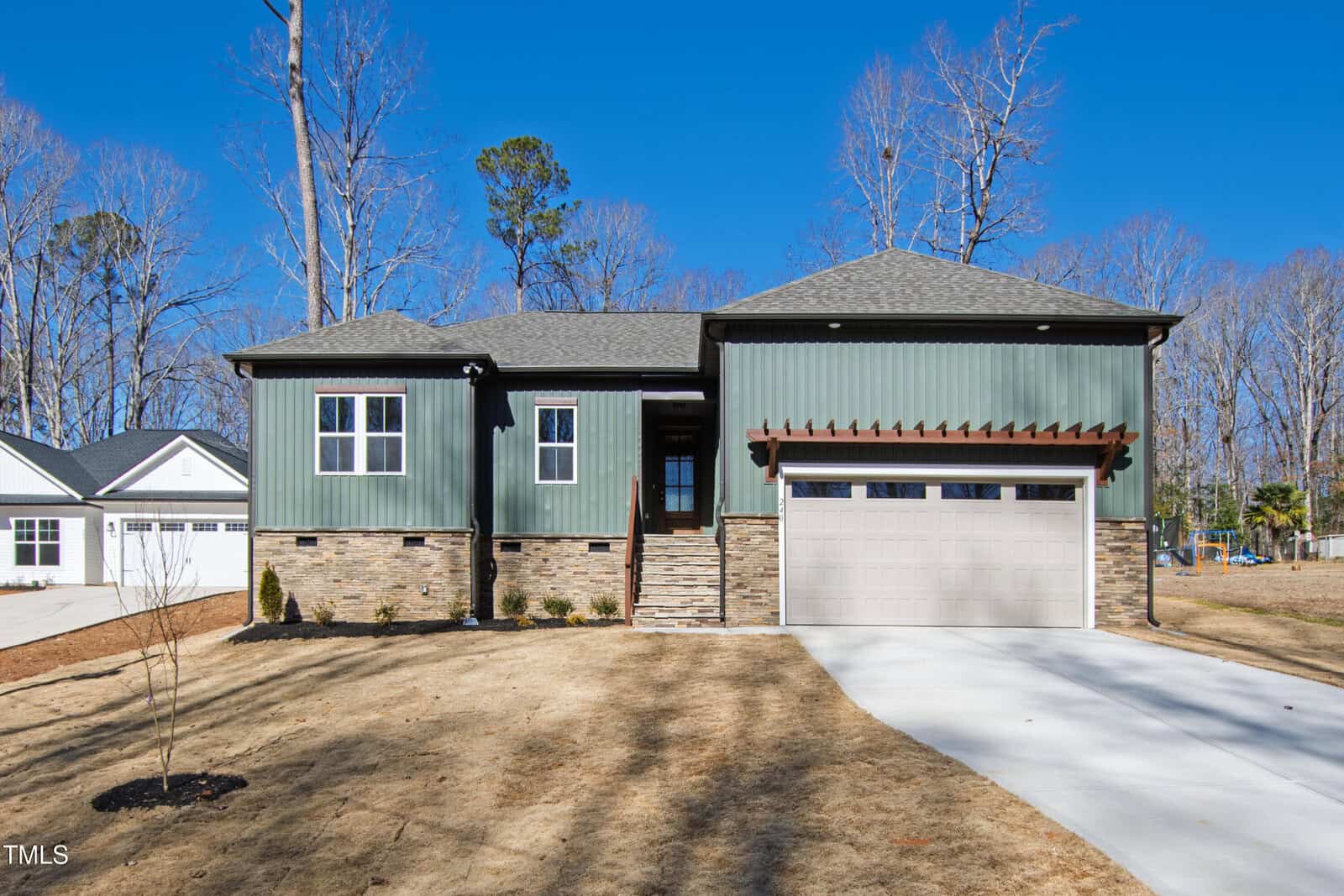 A modern two-story house with green siding and stone accents, featuring a double garage, a small front porch, and a wide concrete driveway. The yard is brown with sparse landscaping and leafless trees in the background.