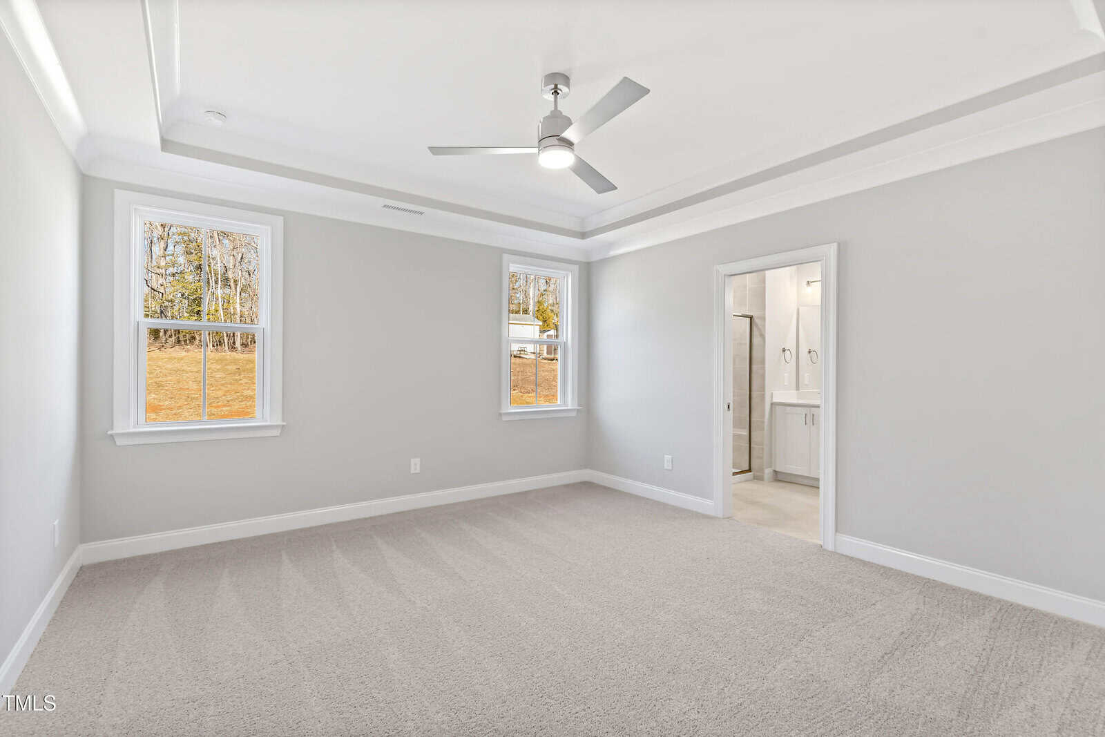 A bright, empty bedroom with light gray walls, beige carpet, a tray ceiling with a ceiling fan, two windows, and an open doorway leading to a bathroom.