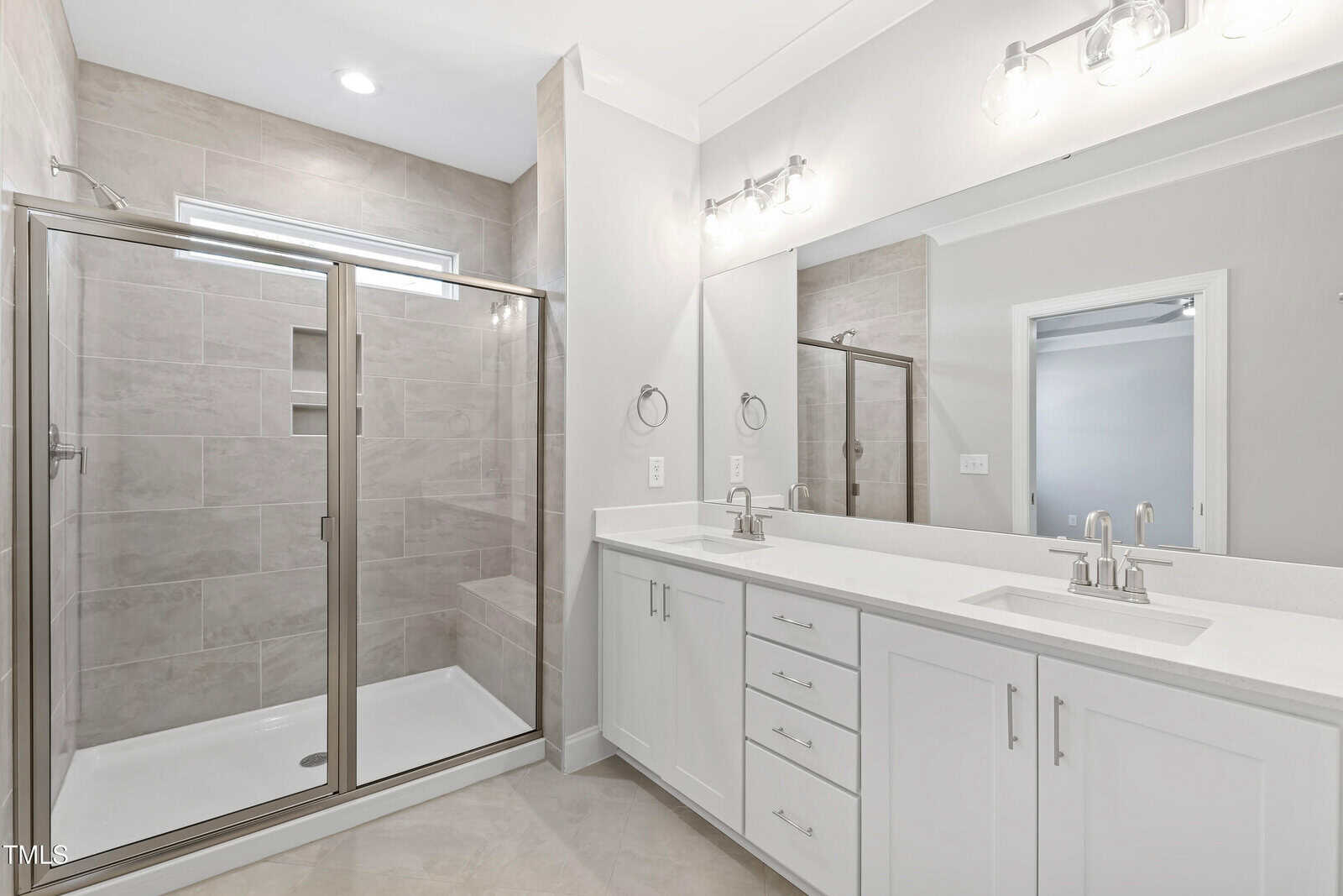 Modern bathroom with a glass shower enclosure, beige tile walls, double sink white vanity with silver fixtures, large mirror, and bright overhead lighting. Door and towel rings visible on the right side.