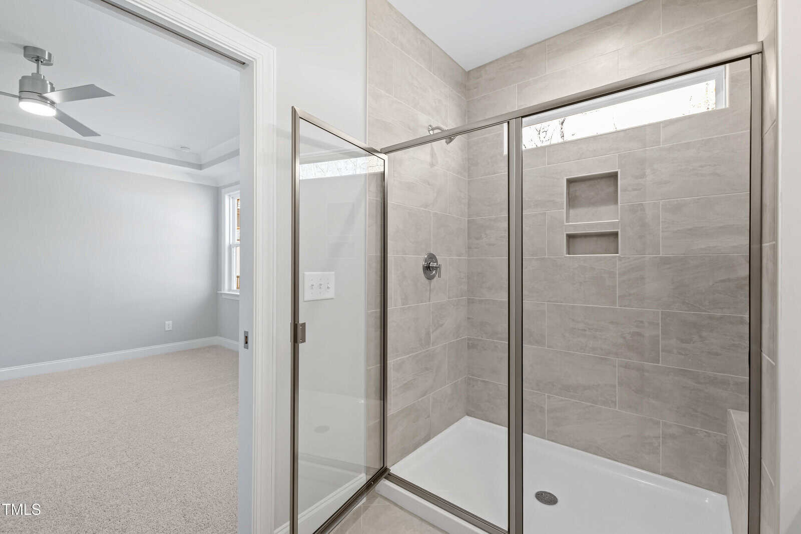 Modern bathroom with a walk-in glass shower, beige tiled walls, built-in shelves, and adjacent carpeted bedroom visible through an open doorway.