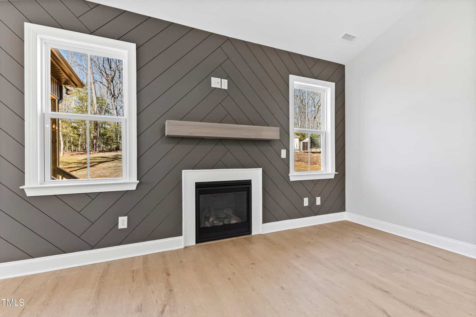 A modern, empty room features light wood flooring, a dark gray chevron accent wall with a linear fireplace, floating mantel, and two windows showing an outdoor view with grass and trees.