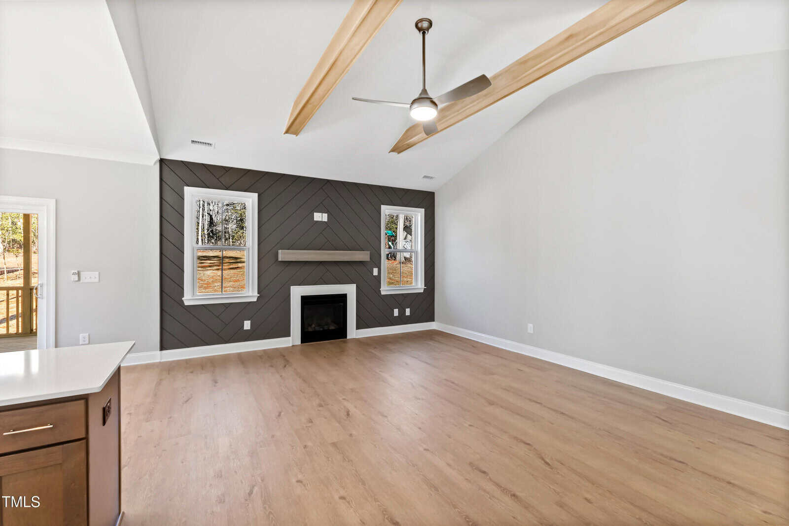 A spacious, empty living room with light wood floors, a vaulted ceiling with wooden beams, a ceiling fan, two windows, a black accent wall with a horizontal pattern, a fireplace, and a floating mantle.