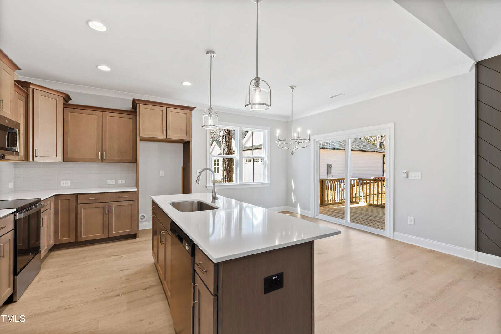 A modern kitchen with light wood cabinets, stainless steel appliances, a large island with a sink, pendant lights, and an adjacent dining area. Sliding glass doors open to a wooden deck outside.