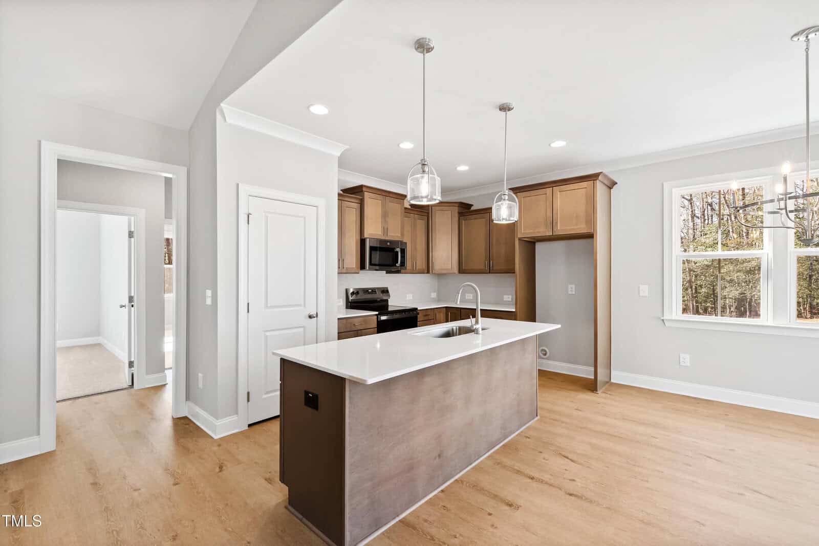 A modern kitchen with wood cabinets, white countertops, stainless steel appliances, pendant lights over a center island, and light wood flooring. Nearby, there is a door, a window, and an open entrance to another room.