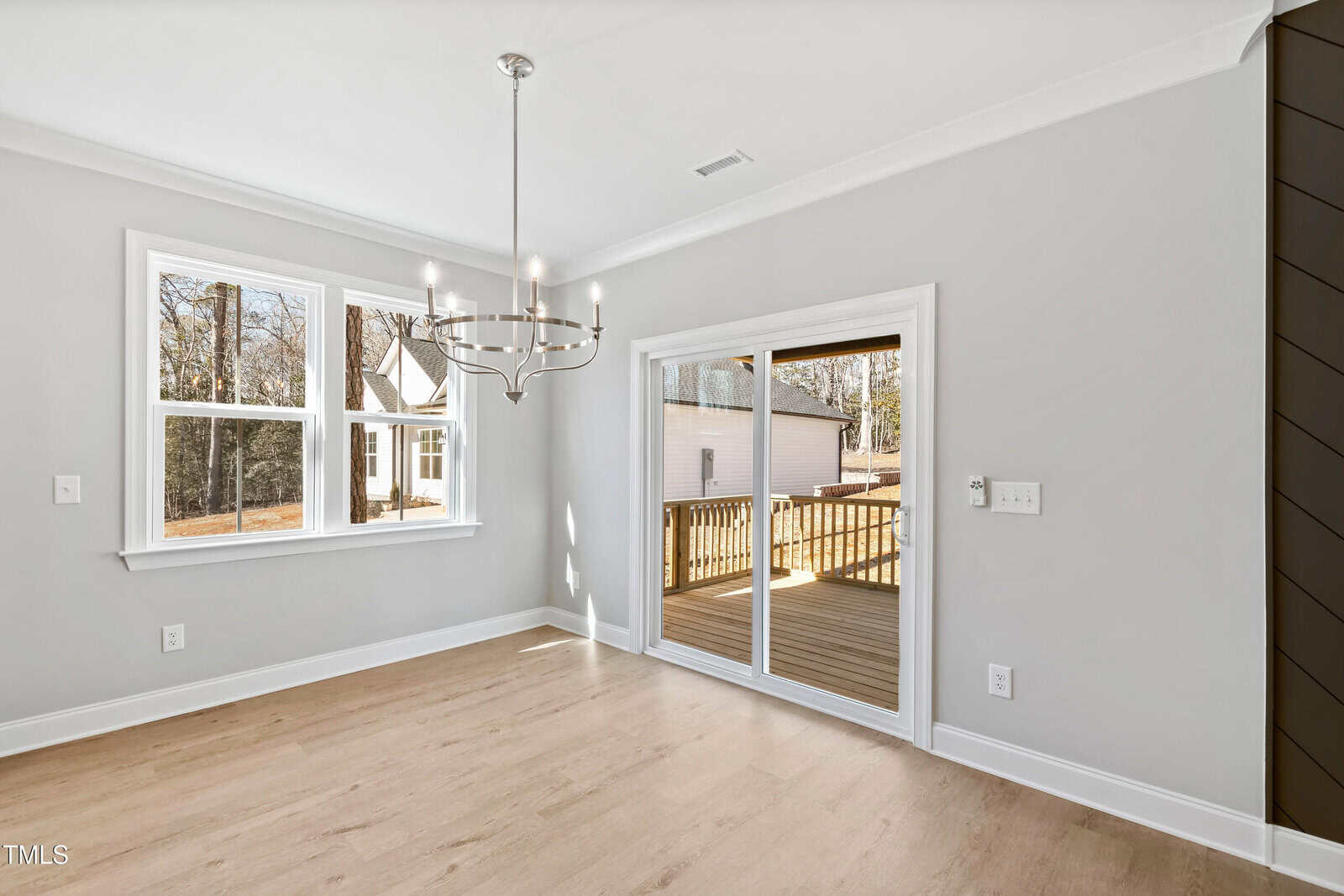 A bright, empty room with light wood floors, gray walls, a modern chandelier, two windows, and a sliding glass door leading to a wooden deck outside.